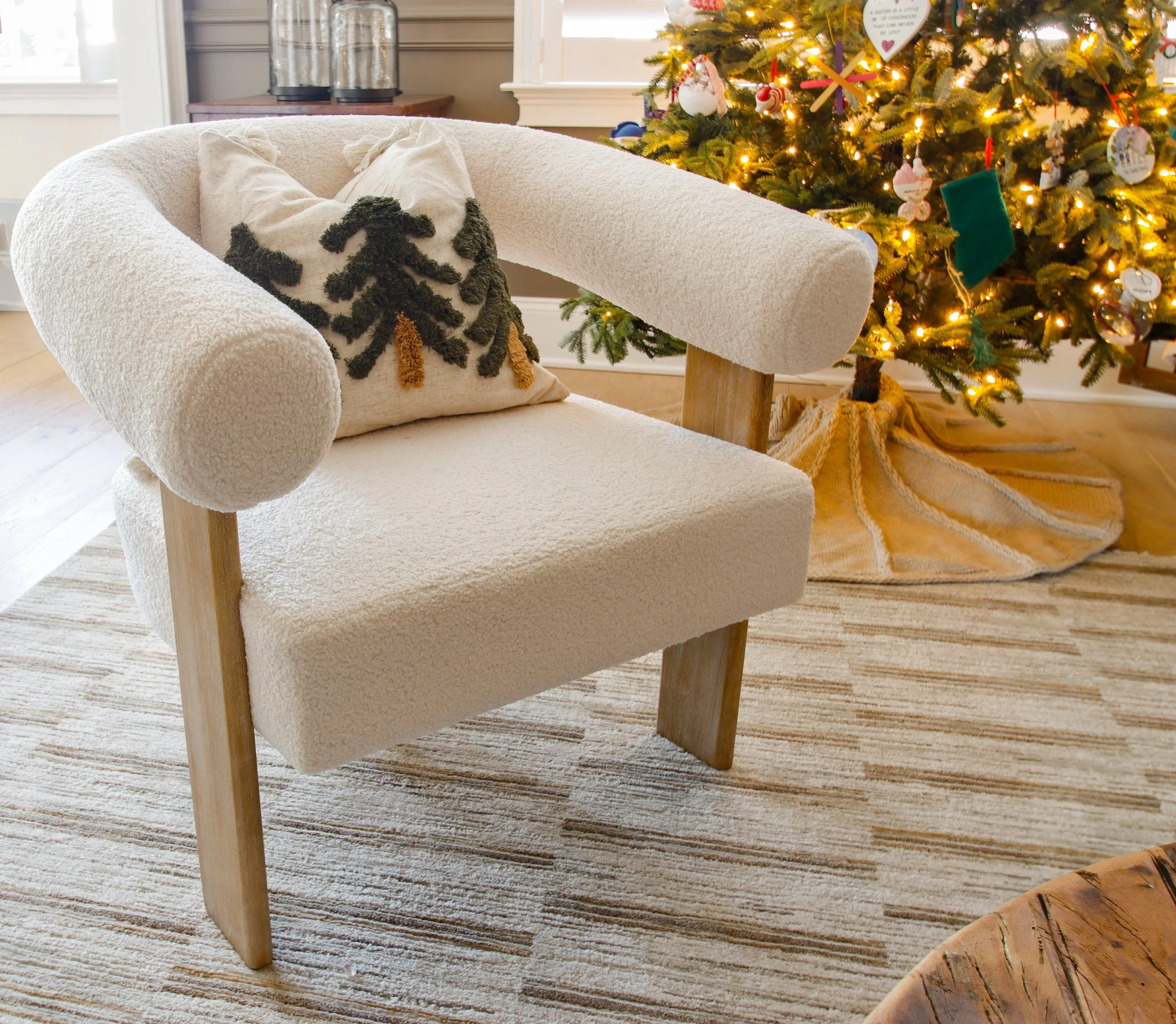 An off-white cozy armchair with a pillow depicting a Christmas tree, situated next to a decorated Christmas tree with lights and ornaments in a living room.