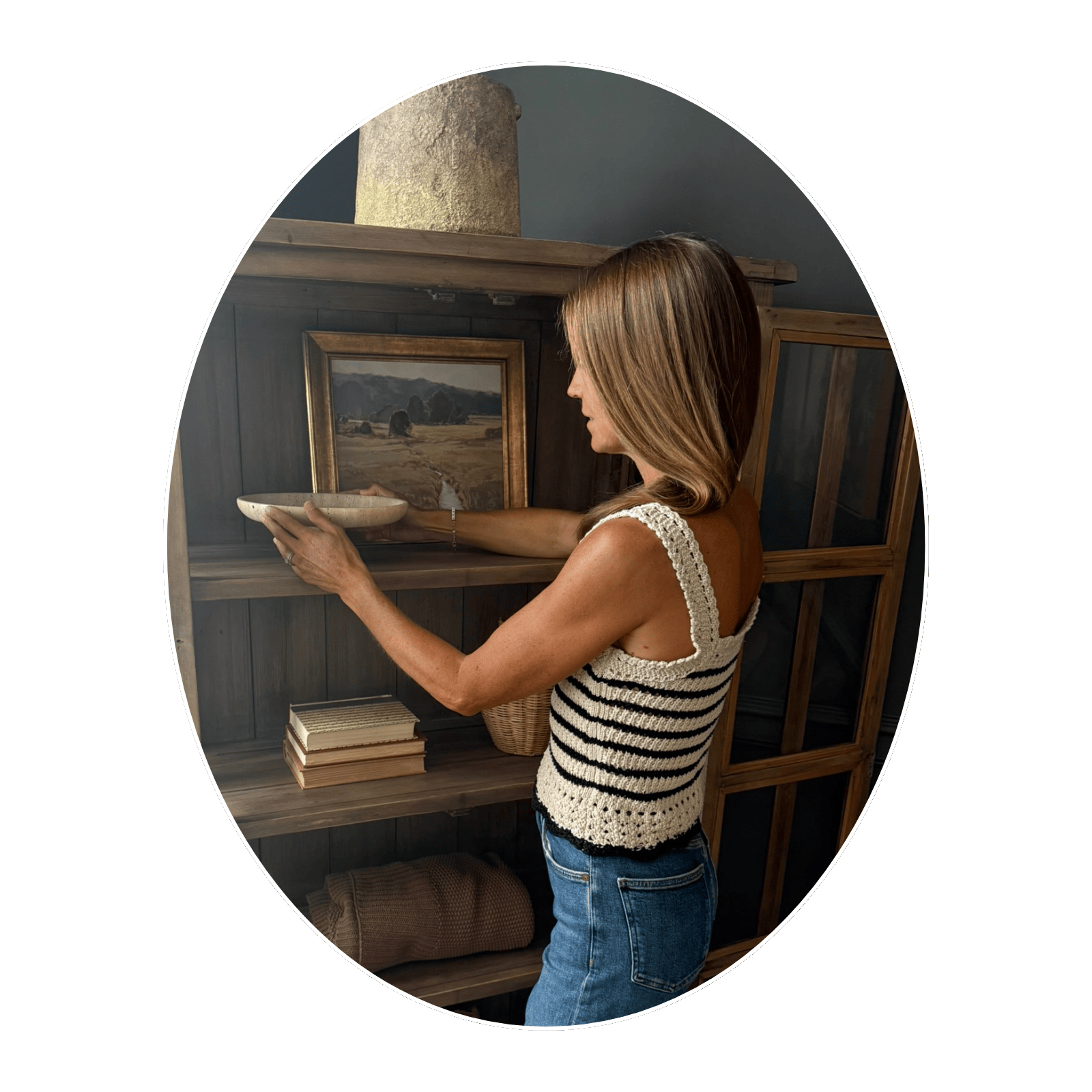 A woman with shoulder-length hair wearing a sleeveless crocheted top and jeans is placing a ceramic bowl on a wooden shelf. Behind her, there are framed landscape paintings and a stack of books.