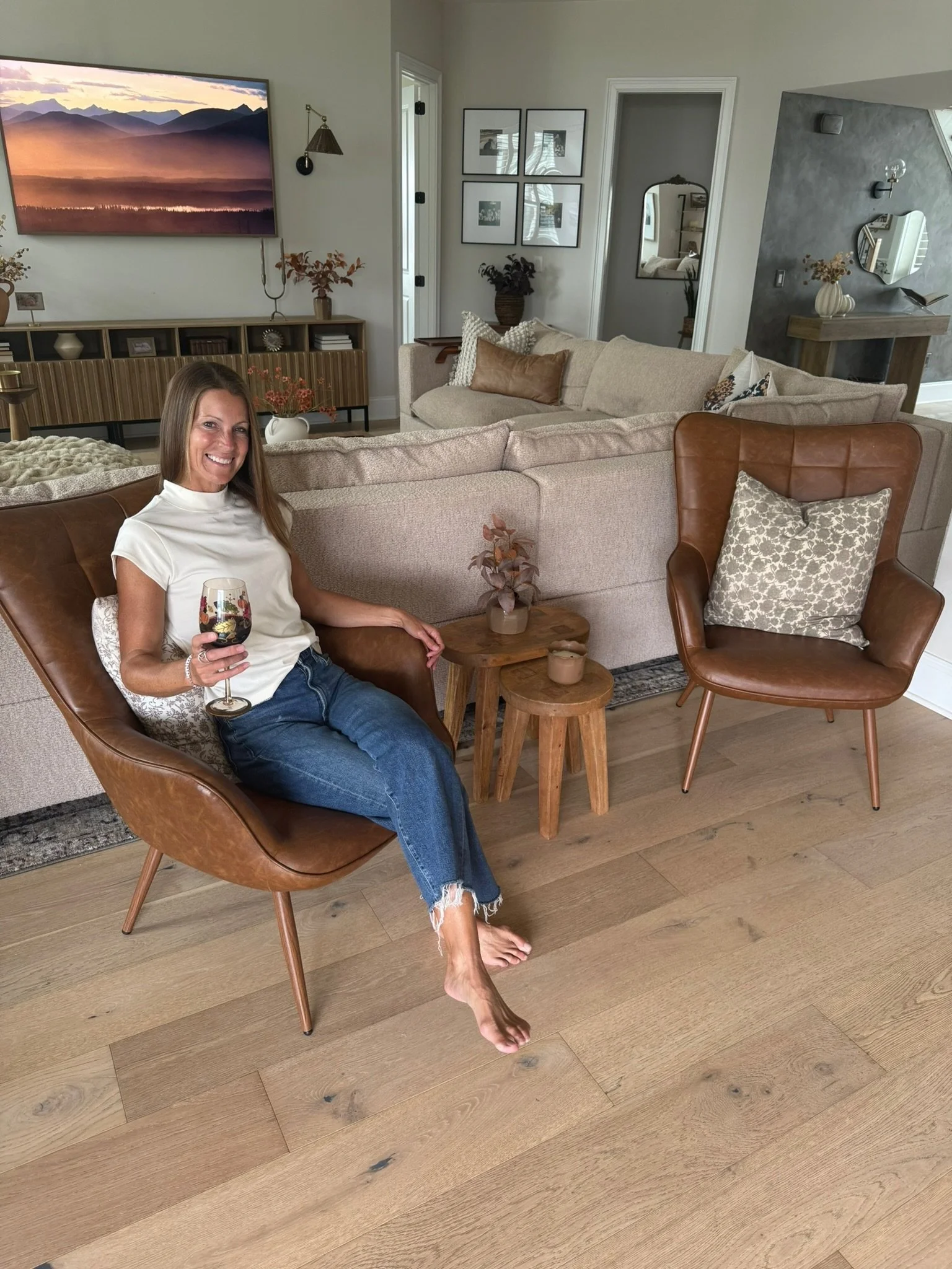 A woman sitting on a brown leather chair holding a glass of red wine in a modern living room with a beige sofa, wooden side tables, a large wall-mounted TV, and various decorative items.