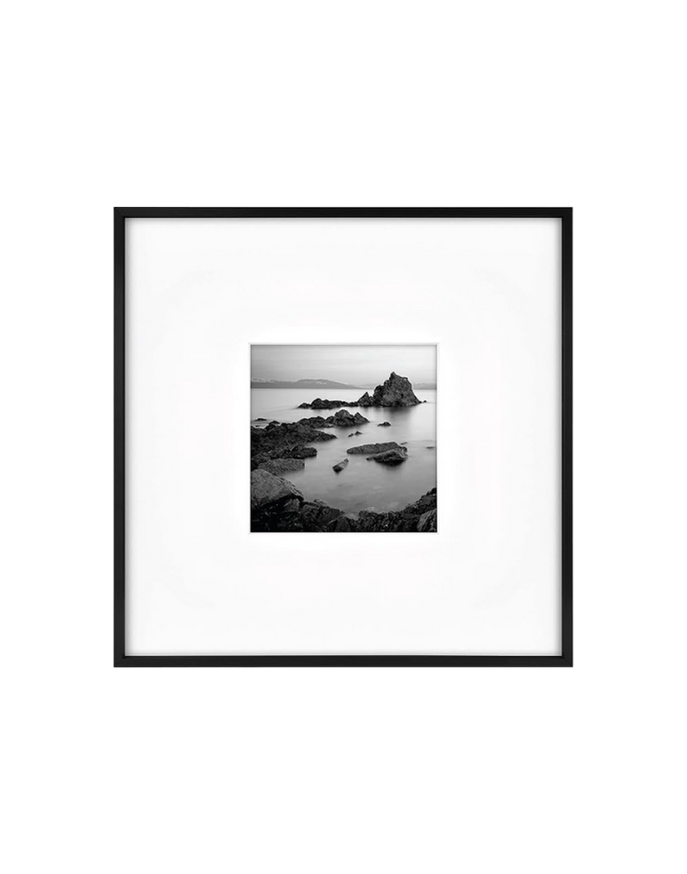 Black and white photograph of a rocky shoreline with a prominent rock formation in the water, displayed in a black frame with a white matte, centered on a black background.