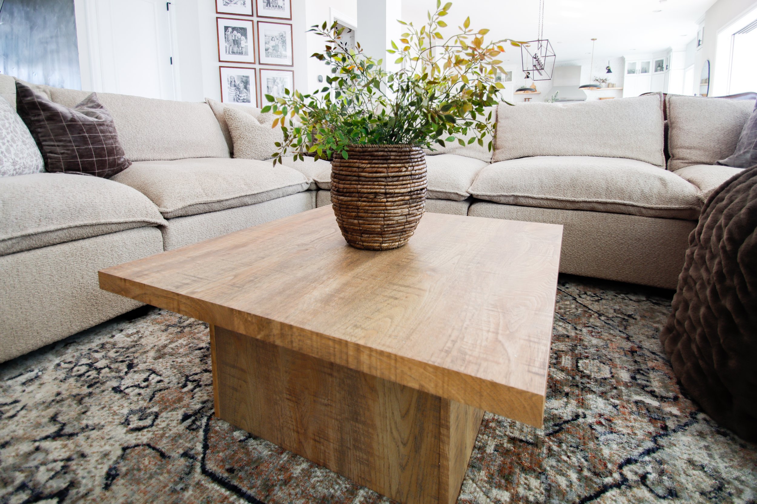 Living room with a beige sectional sofa, a wooden coffee table with a woven basket-style vase, and a patterned rug.