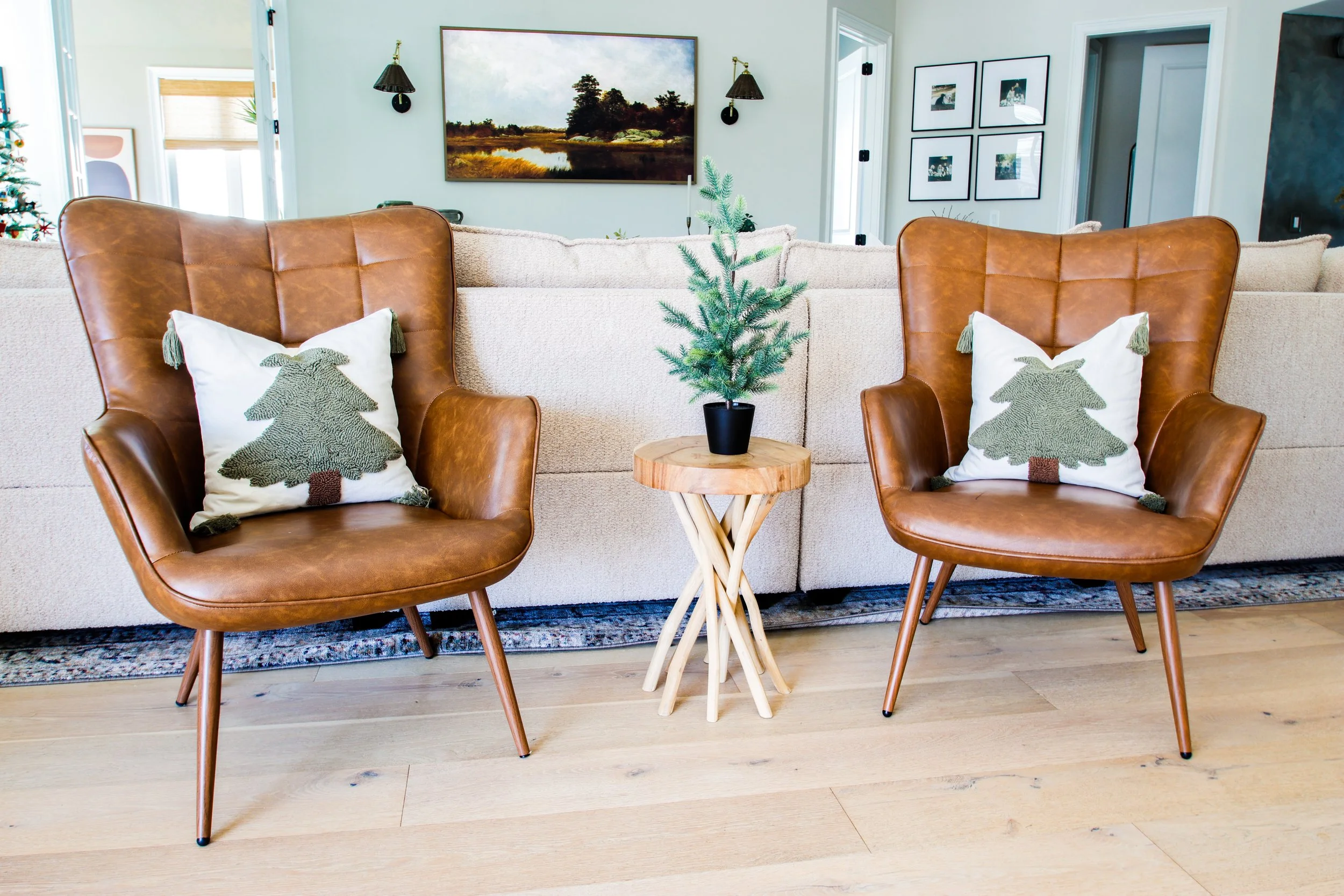 Living room with two brown leather armchairs with Christmas tree pillows, a small wooden side table with a potted pine tree, and a beige sofa in the background, decorated for the holidays.