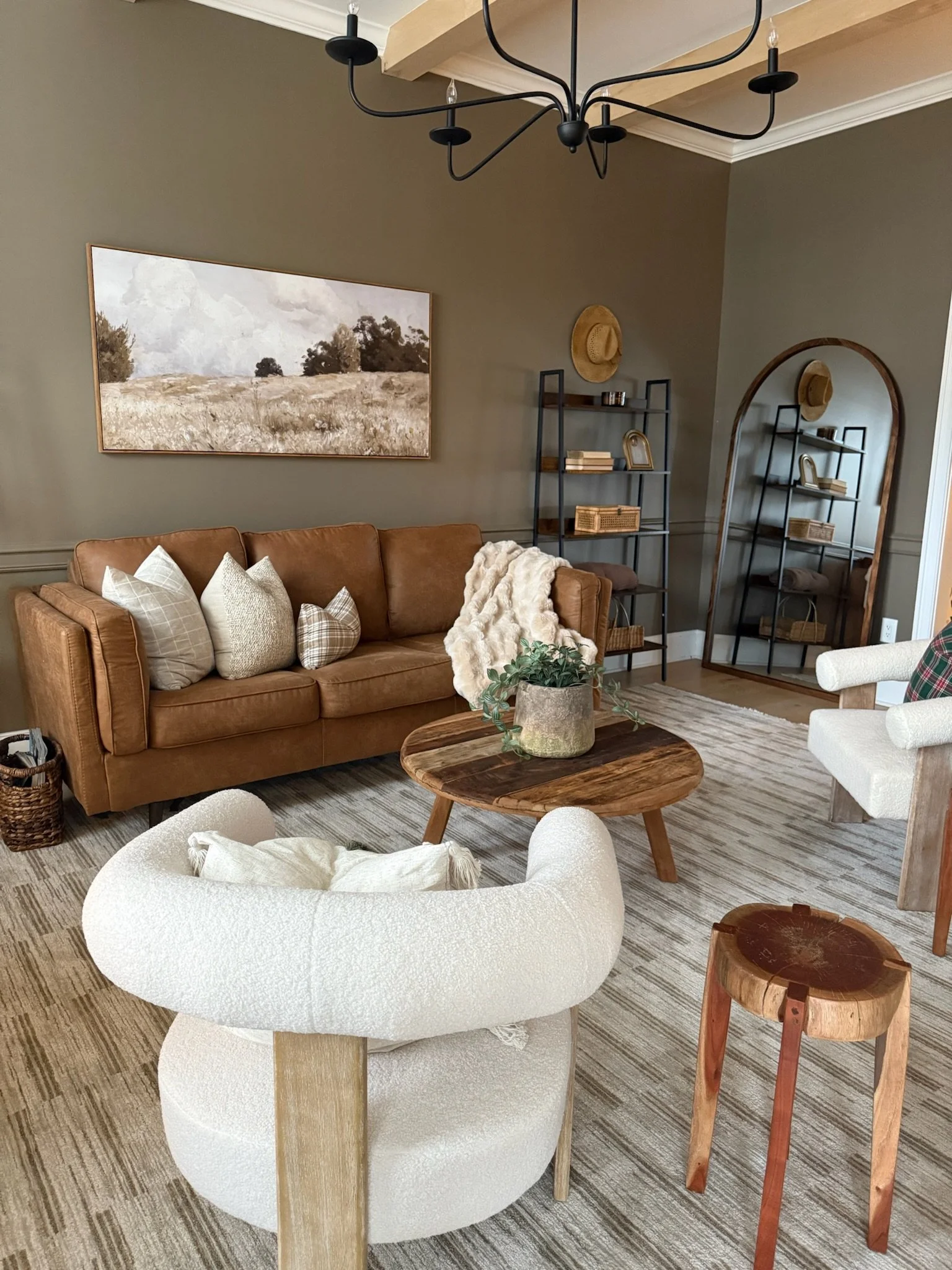 Cozy living room with beige sofa, white and plaid throw pillows, and a tan throw blanket. Wooden coffee table with a plant in a stone pot. White textured armchair, small wooden side table. Wall art, shelves with books and baskets, wall mirror, and a 