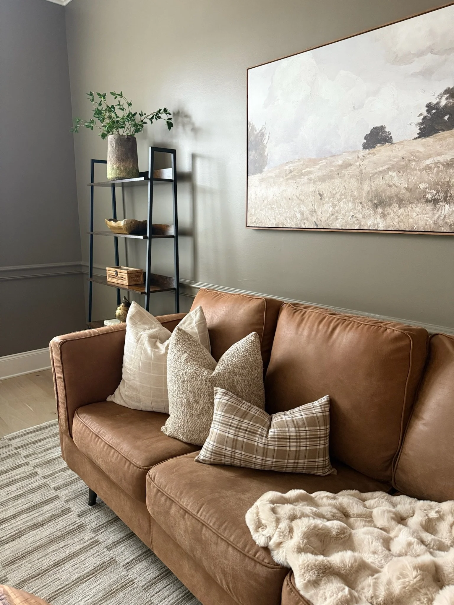 A cozy living room with a brown leather sofa adorned with beige and plaid throw pillows. A soft cream-colored blanket is draped over the armrest. Behind the sofa, a large landscape artwork hangs on the light gray wall. To the left, a black metal and 