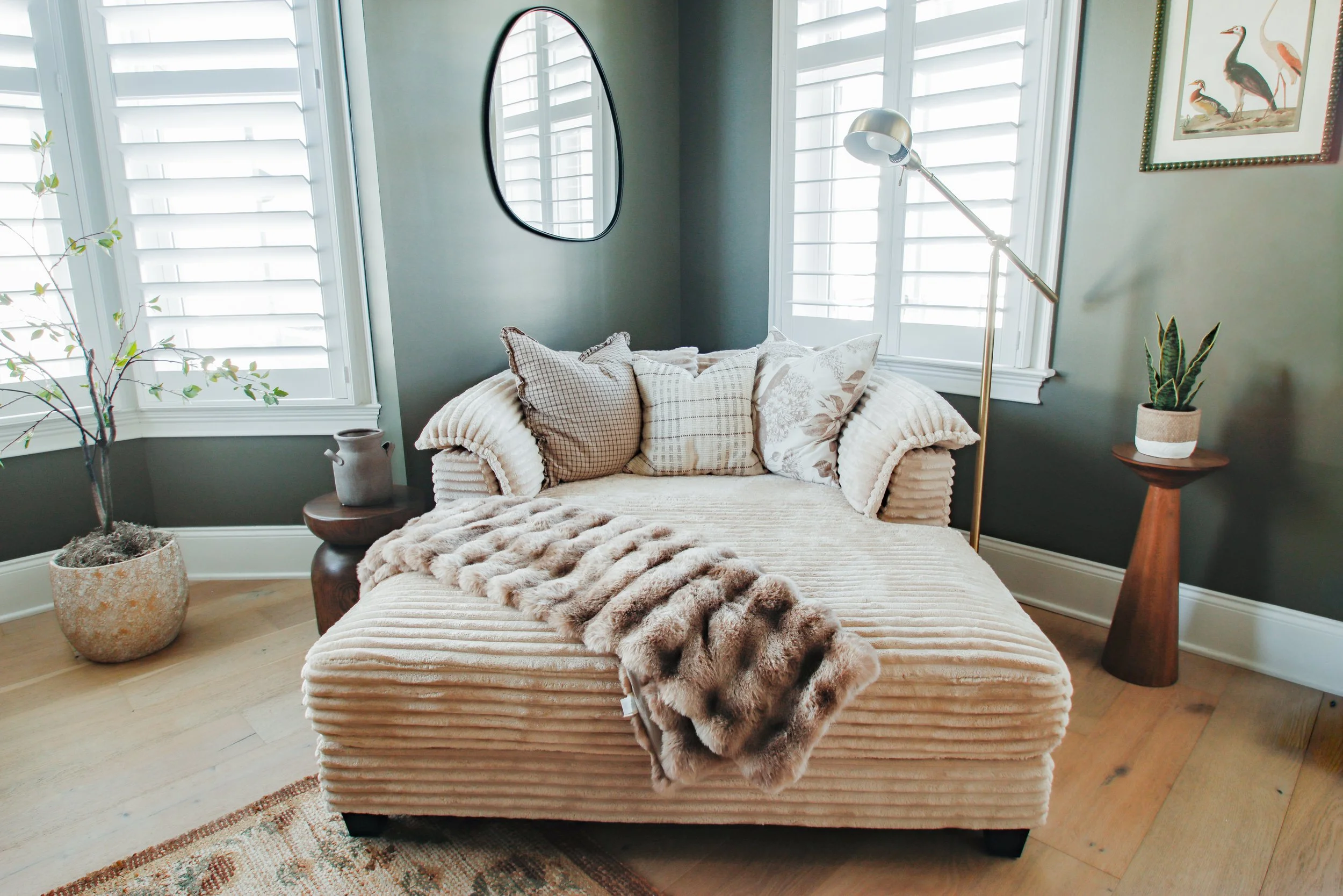 Living room corner featuring a beige upholstered sofa with textured throw blanket and assorted pillows, flanked by a potted plant and decorative vase, with shutters on the windows and a floor lamp.
