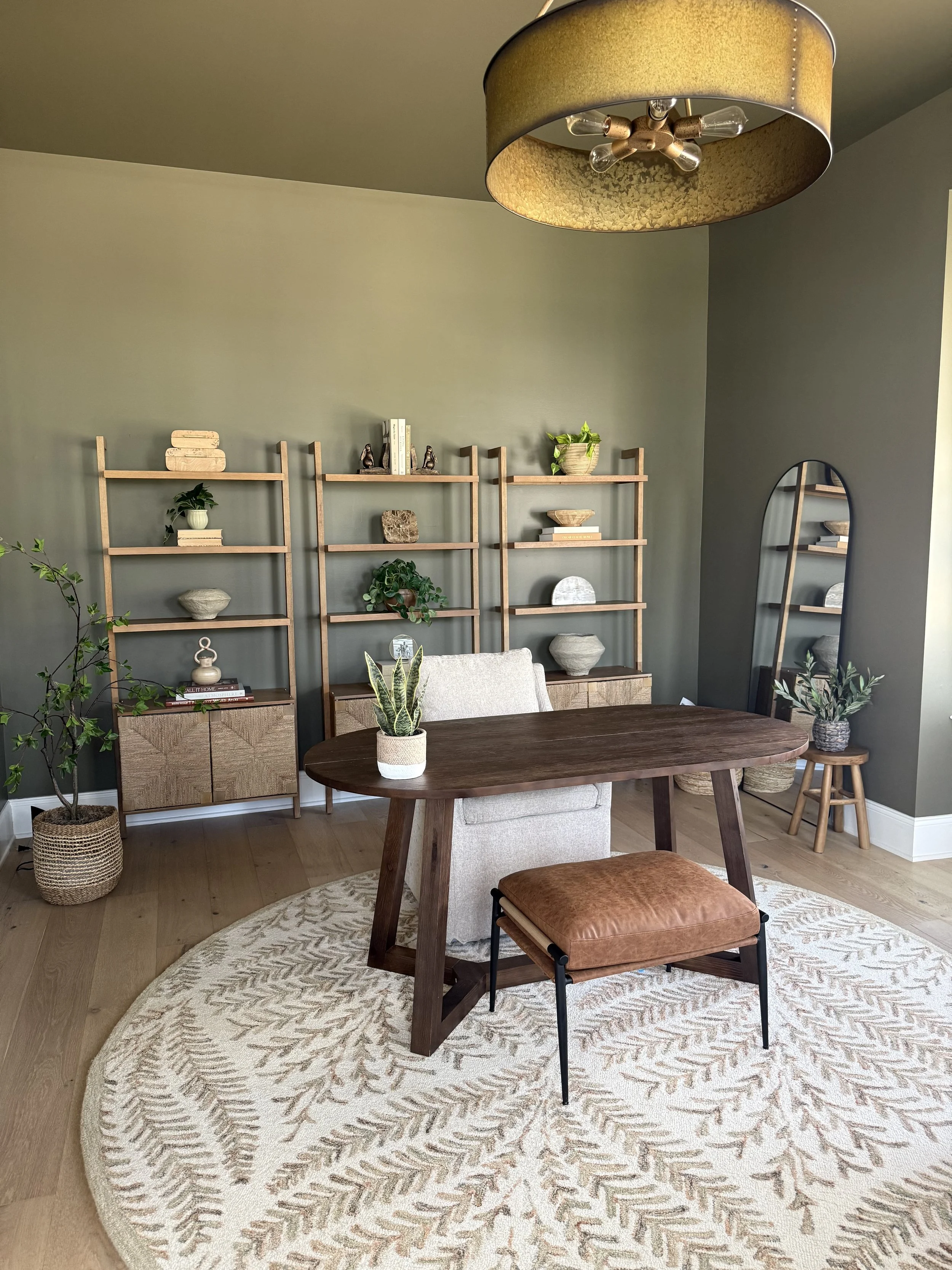 Modern home office with a large wooden desk, a white cushioned chair, a tall mirror, and a wooden bookshelf with decorative items and plants on a light green wall, with a patterned rug on the floor.