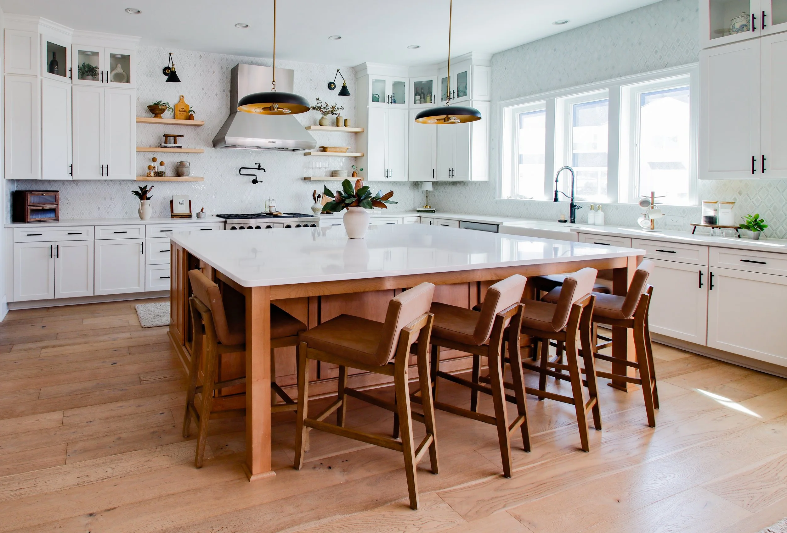Bright kitchen with white cabinets, a large island with a white countertop and wooden base, six brown chairs around the island, and natural light from multiple windows.