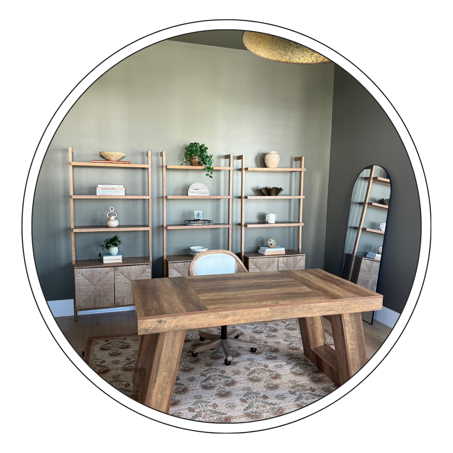 Modern home office with a large wooden desk, a white cushioned chair, a tall mirror, and a wooden bookshelf with decorative items and plants on a light green wall, with a patterned rug on the floor.