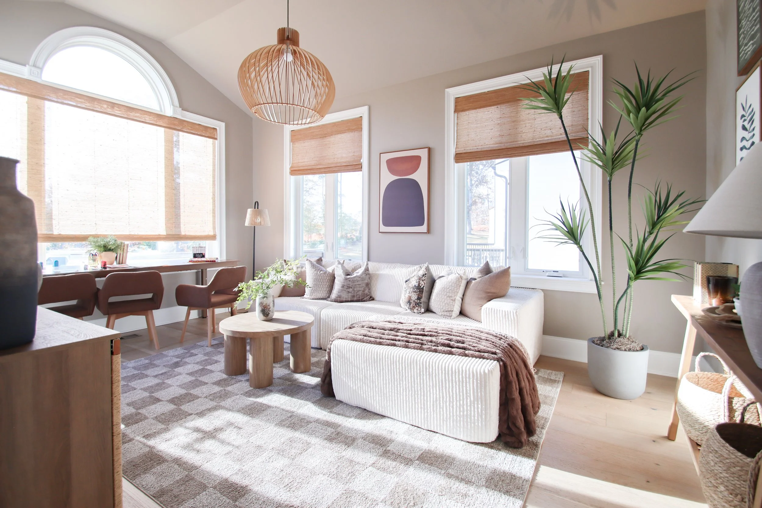 Bright living room with large windows, beige sofa with pillows, round wooden coffee table, potted plants, and modern decor.