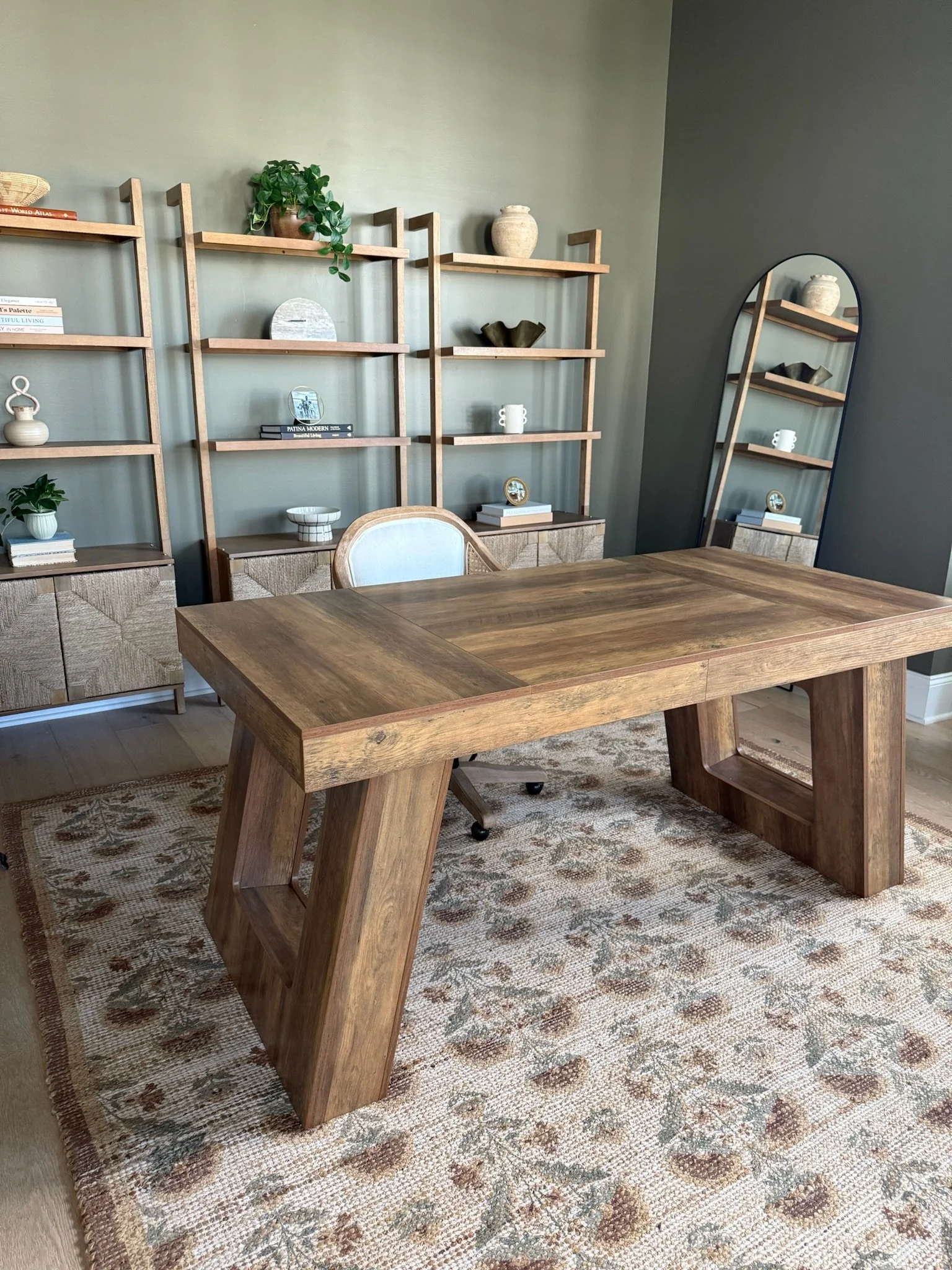 A modern home office with a large wooden desk, a light-colored upholstered chair, a decorative shelf with books, plants, and vases, a large mirror, and a patterned rug.