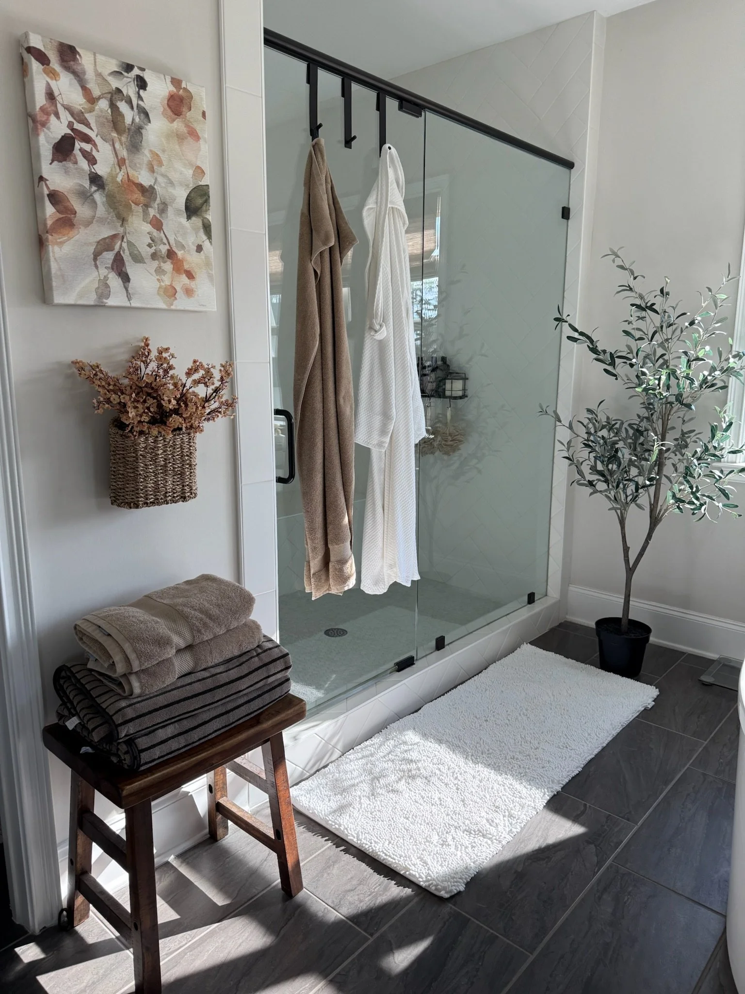 A bathroom with a glass shower door, hanging towels, a small stool with folded towels, a potted plant, and a white bath mat on dark tiled flooring.