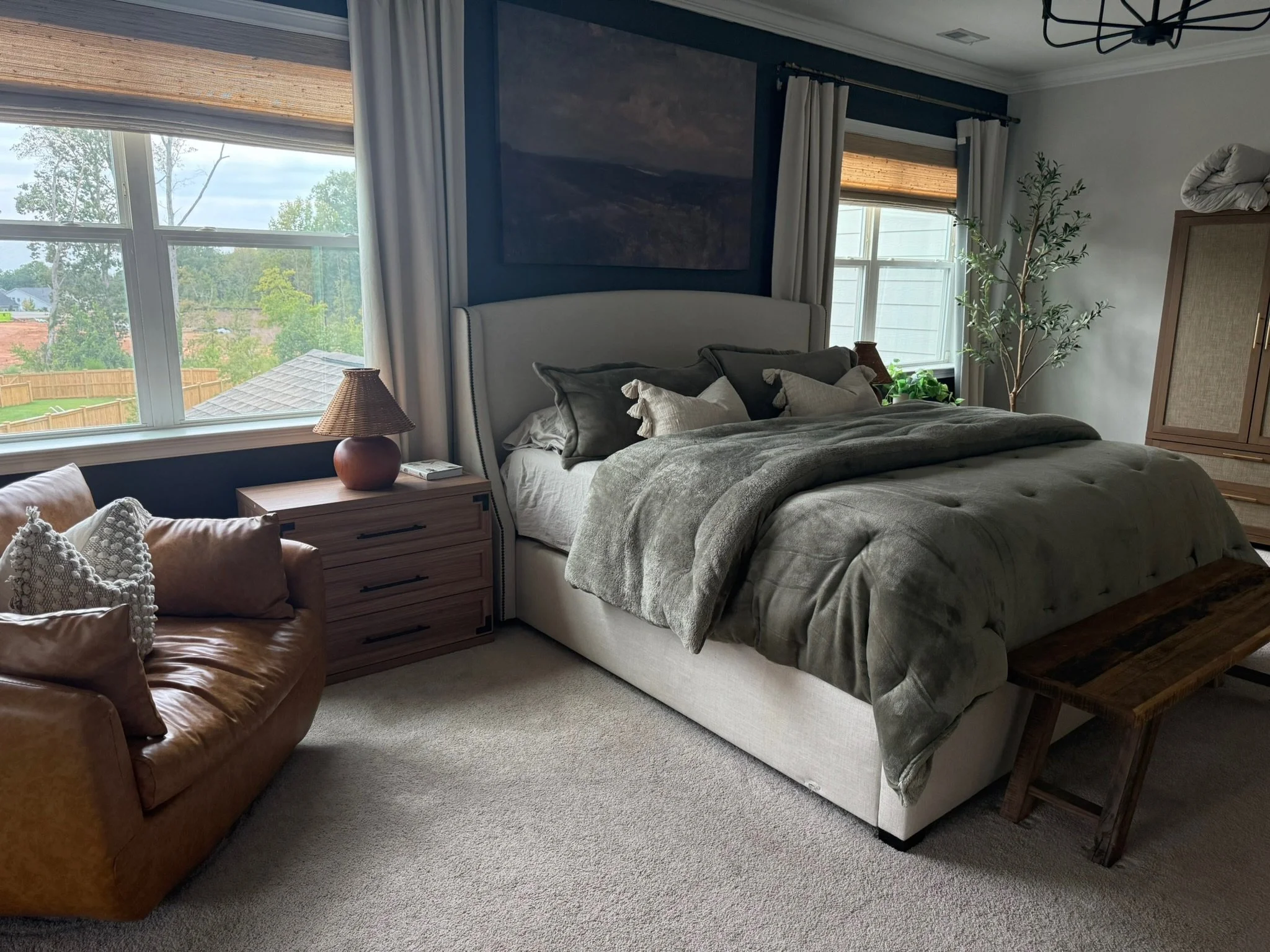 A cozy bedroom with a large bed, window views of a backyard, a side table with a lamp, and a leather armchair with decorative pillows.