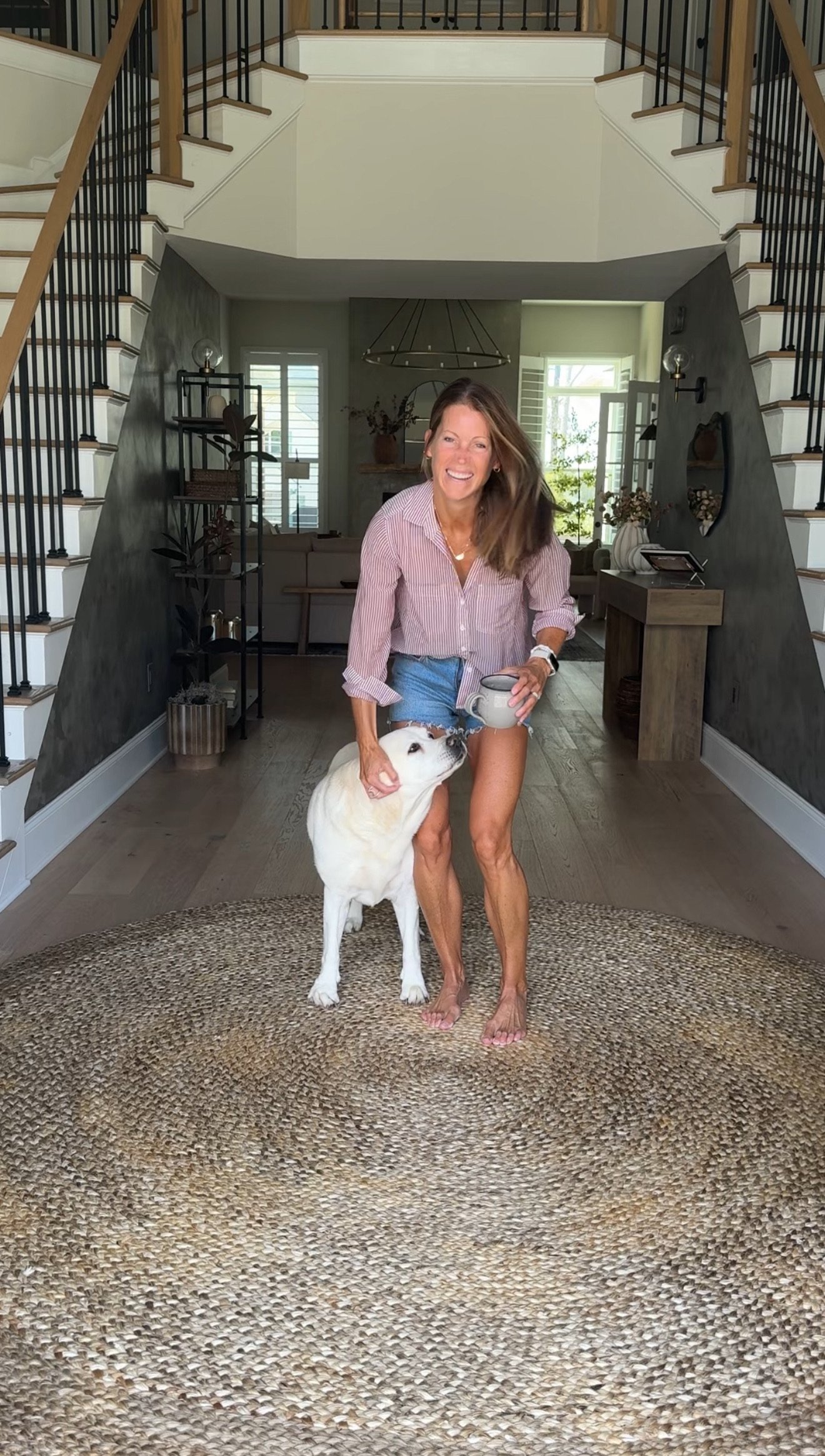 A woman with brown hair, wearing a striped pink shirt and denim shorts, smiling and holding a mug, standing on a circular woven rug indoors with a happy white dog leaning against her.