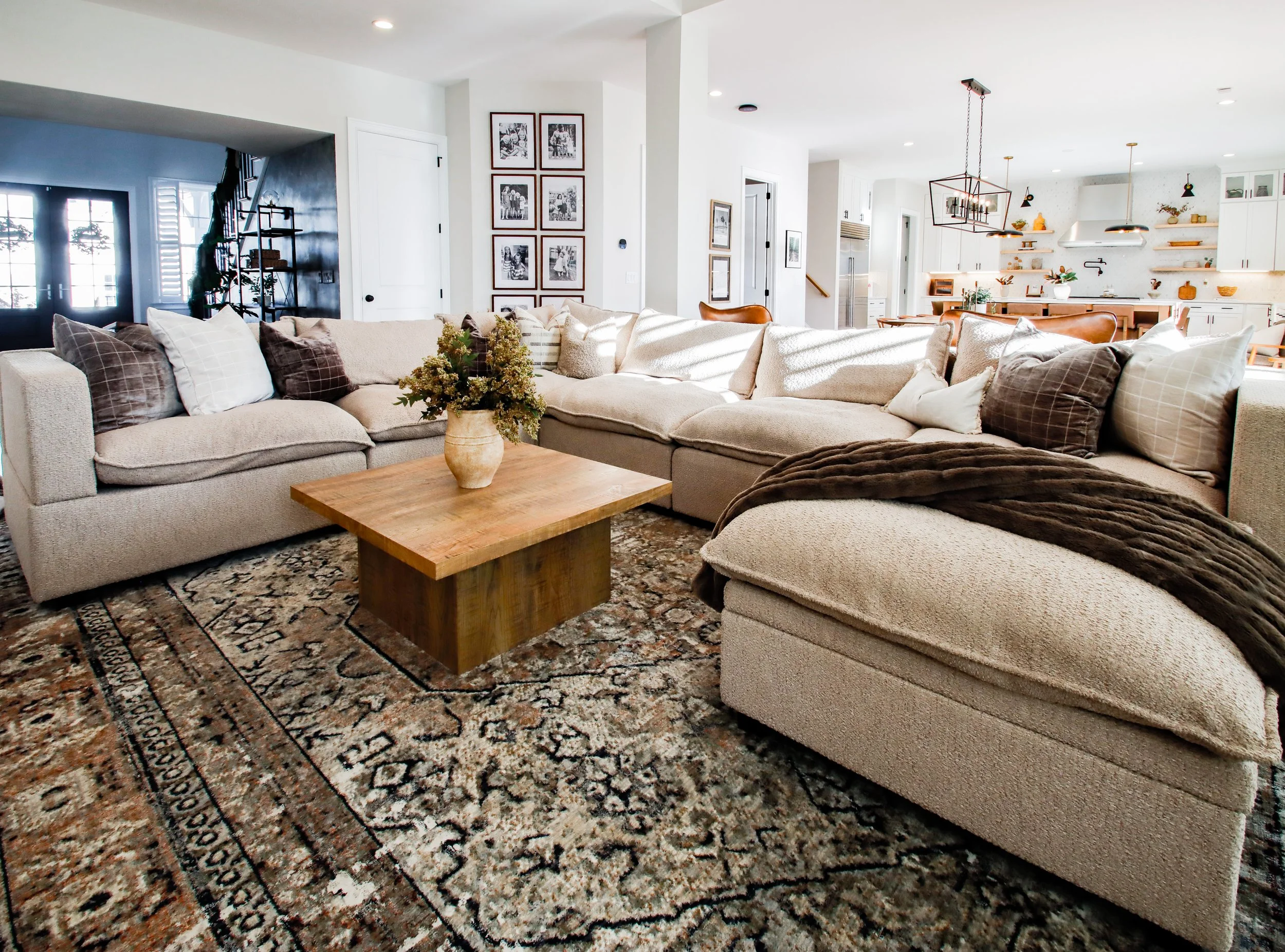 Living room with beige sectional sofa, wooden coffee table with a flower vase, patterned rug, and open kitchen in the background.