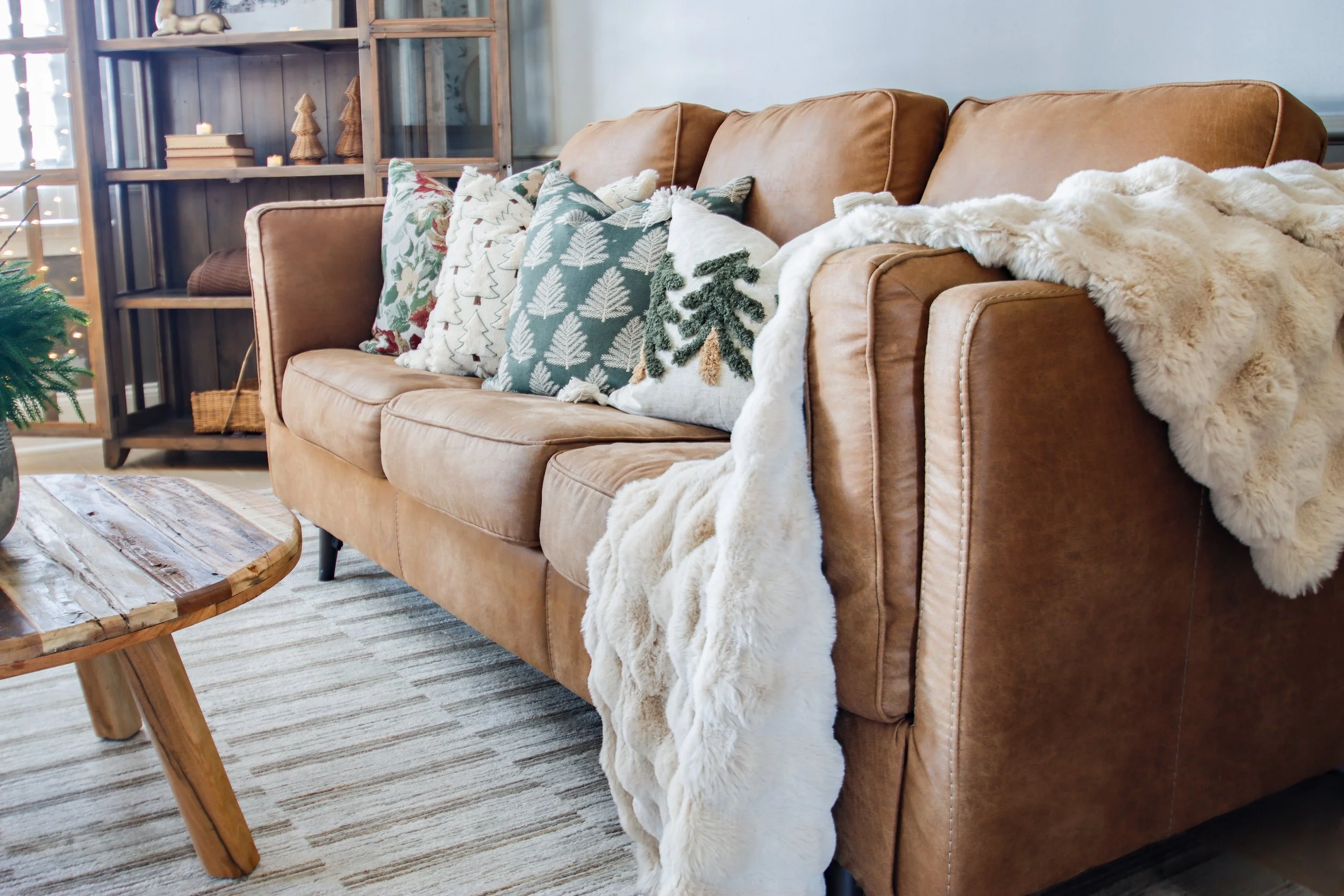 Living room with a brown leather sofa decorated with holiday-themed pillows, a white faux fur throw, a wooden coffee table, a rug, and a bookshelf with candles and Christmas decorations.