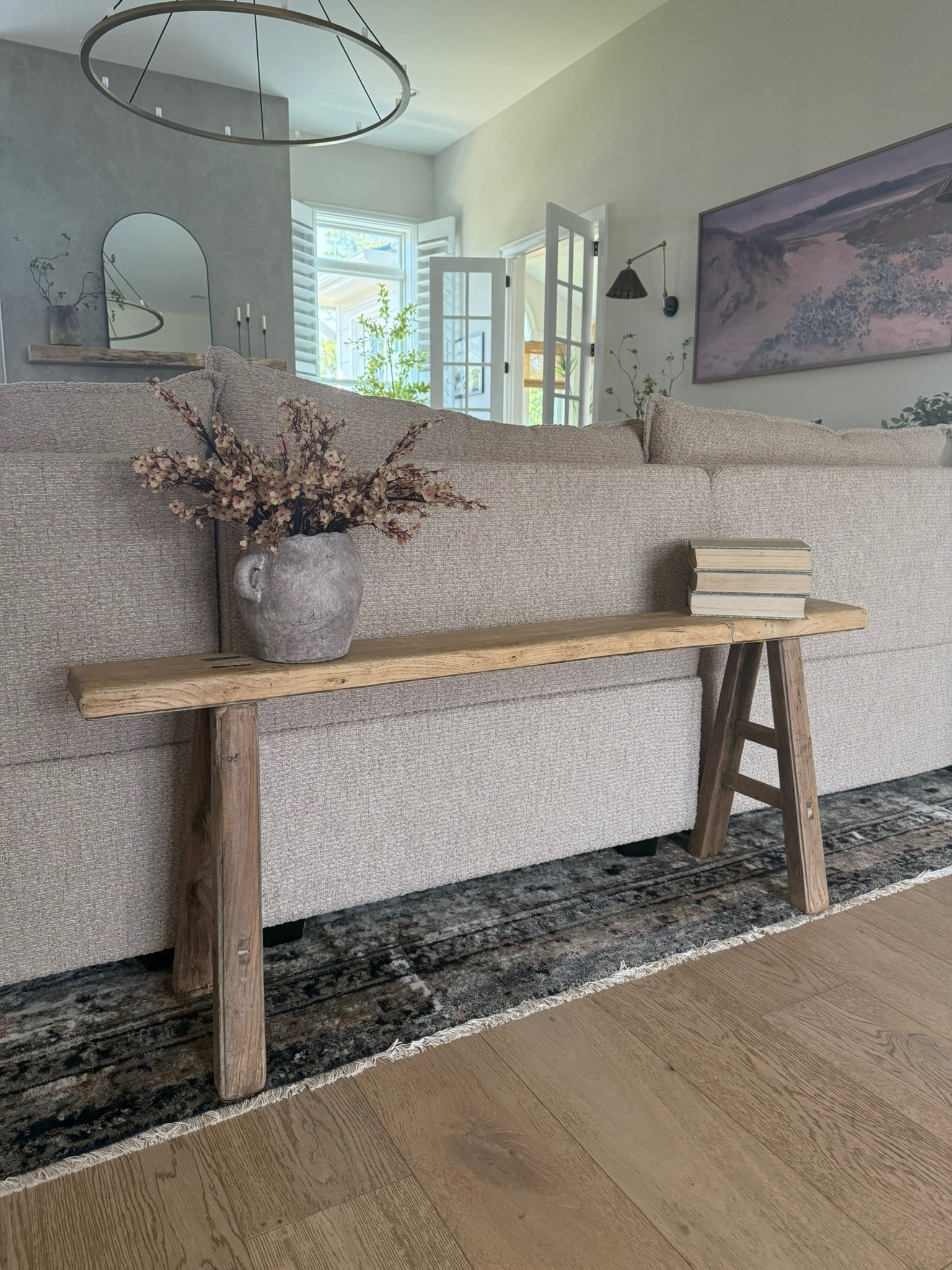 A neutral-colored living room with a beige sofa, wooden side table with a vase of pink blossoms and a stack of books, large window with shutters, and wall art depicting a landscape scene.