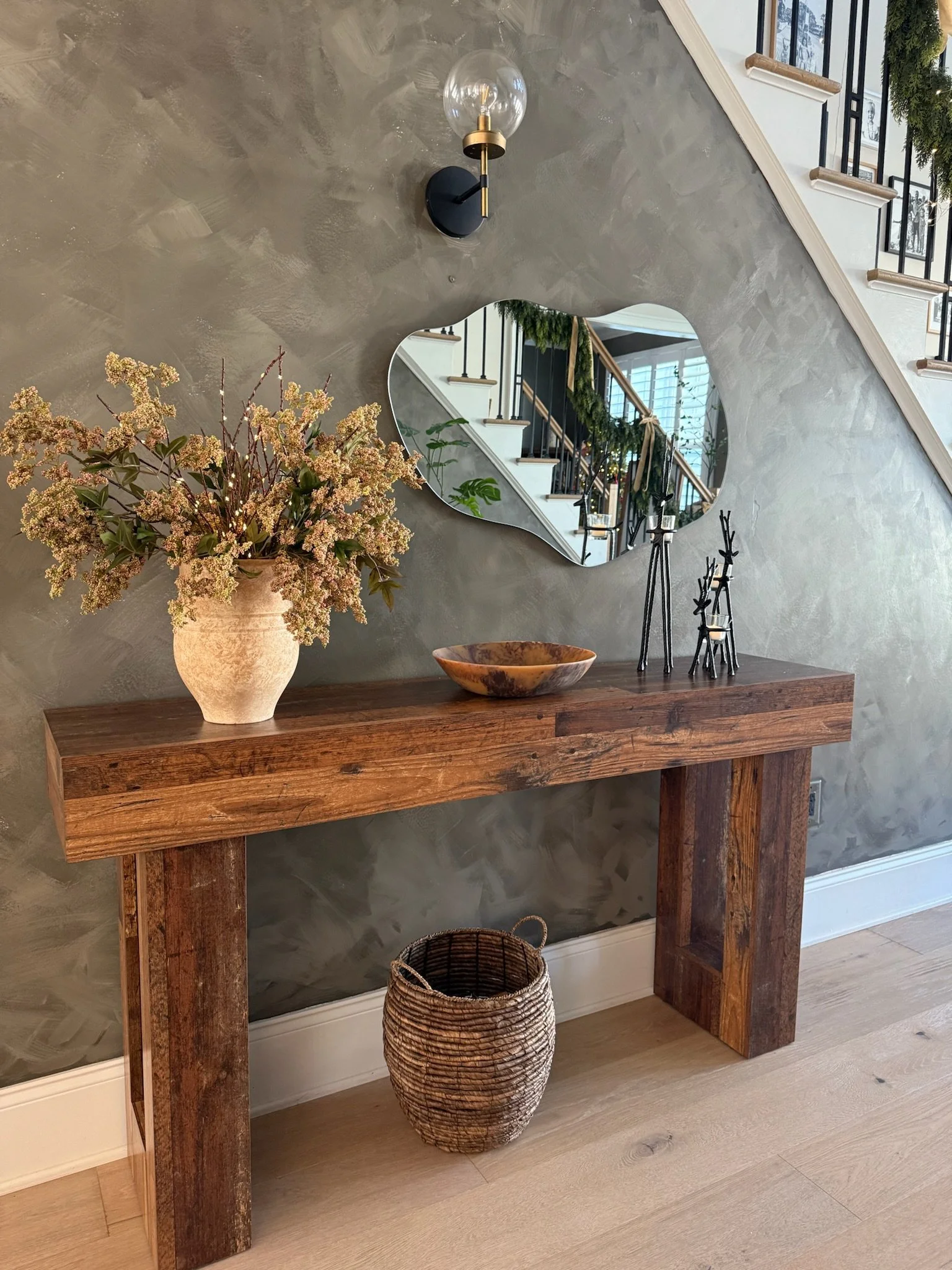 Interior entryway with a rustic wooden console table against a textured gray wall, decorated with a large potted floral arrangement, a wooden bowl, and black metal figurines. A mirror with an organic shape hangs on the wall, reflecting a staircase wi