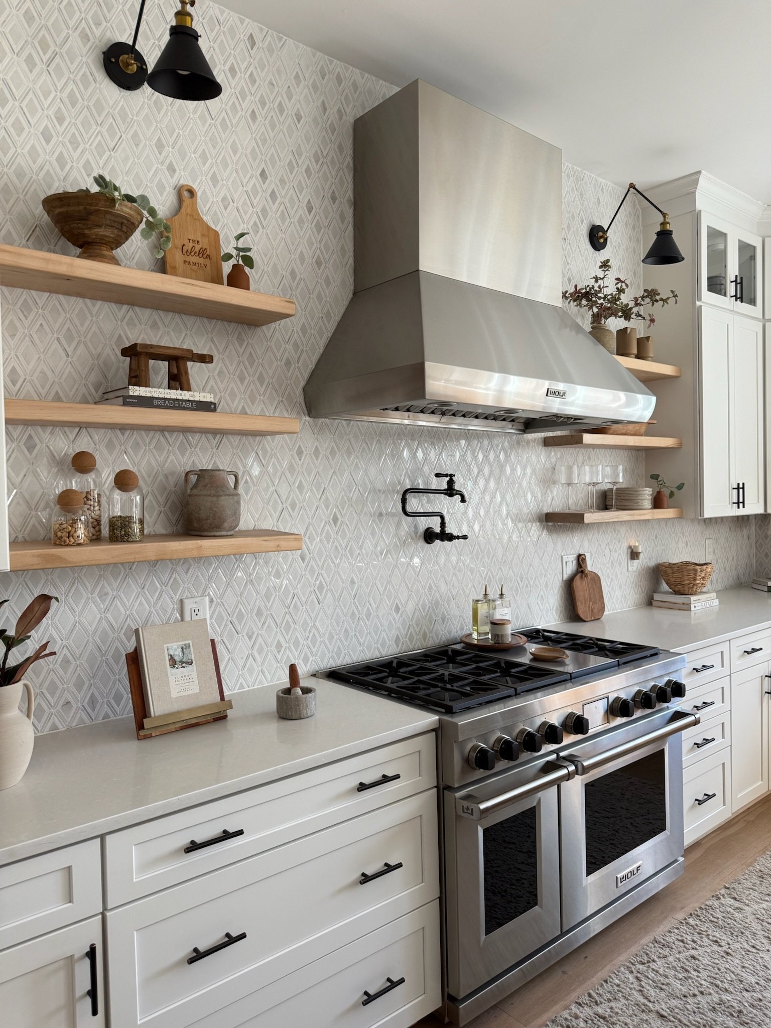A modern kitchen with white cabinets, a stainless steel oven, and open wooden shelves with decorative items. There are black wall-mounted lamps and a textured backsplash.