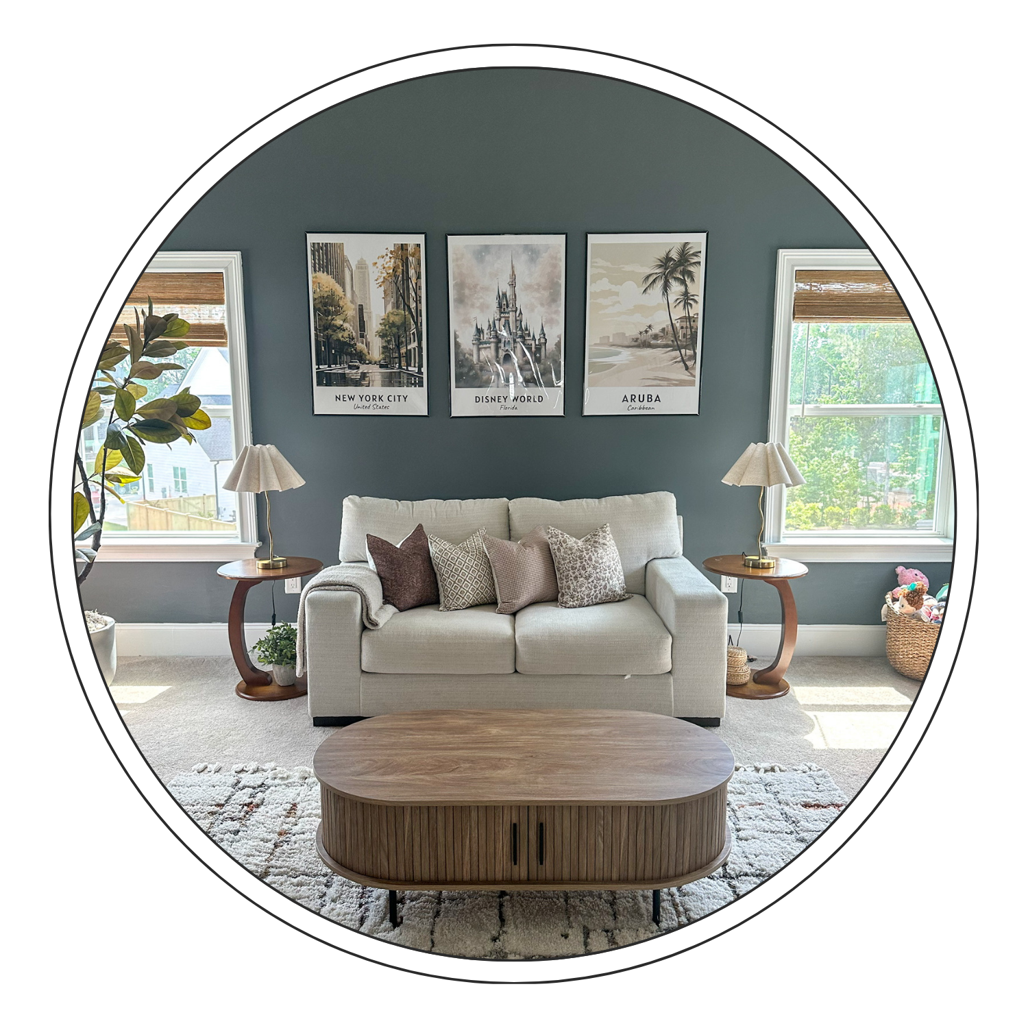 Living room with a beige sofa, decorative pillows, two side tables with lamps, wall art depicting New York City, Disney World, and Aruba, and two windows with blinds overlooking greenery.