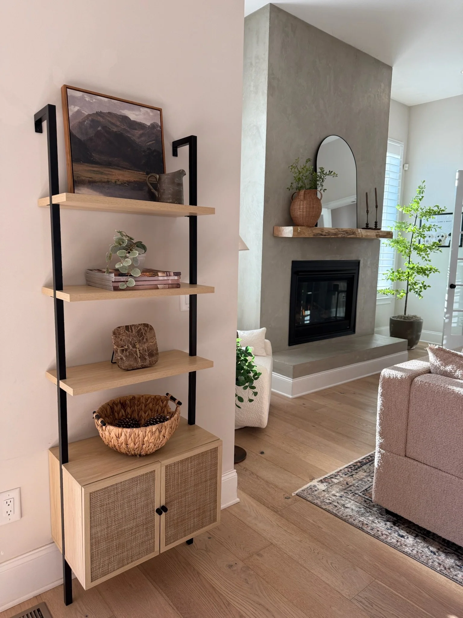 Living room with a beige sofa, a wooden and black metal shelf with decorative items including a landscape painting, books, and pottery, a fireplace with a stone mantel, a large mirror, plants, and a window with white shutters, on wooden flooring.