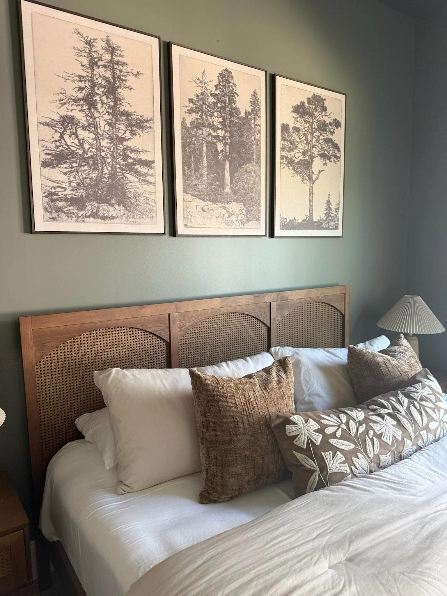 A bedroom with a wooden headboard, beige decorative pillows, dark bedside lamp, and a landscape painting on a dark green wall.