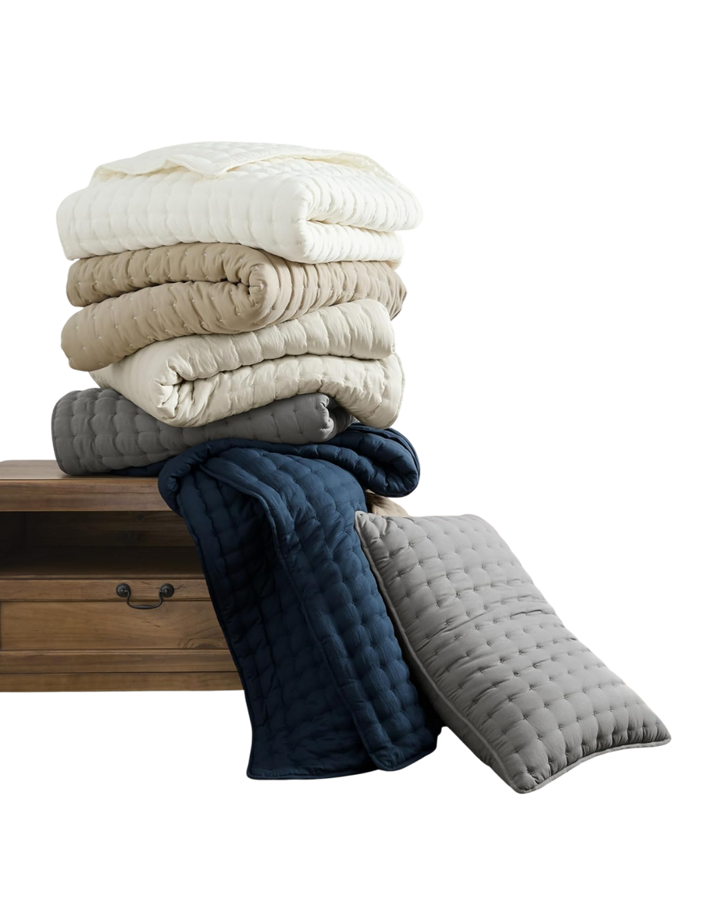 A stack of folded quilts in various neutral colors on a wooden nightstand with additional quilts draped over the edge.