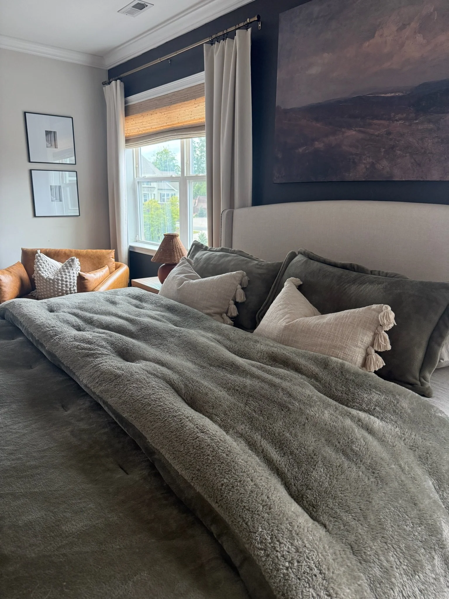 A cozy bedroom with a bed featuring multiple pillows and a plush blanket. There is a window with white curtains, a tan sitting area with cushions, and framed artwork on the walls. The room has dark and light contrasting decor.