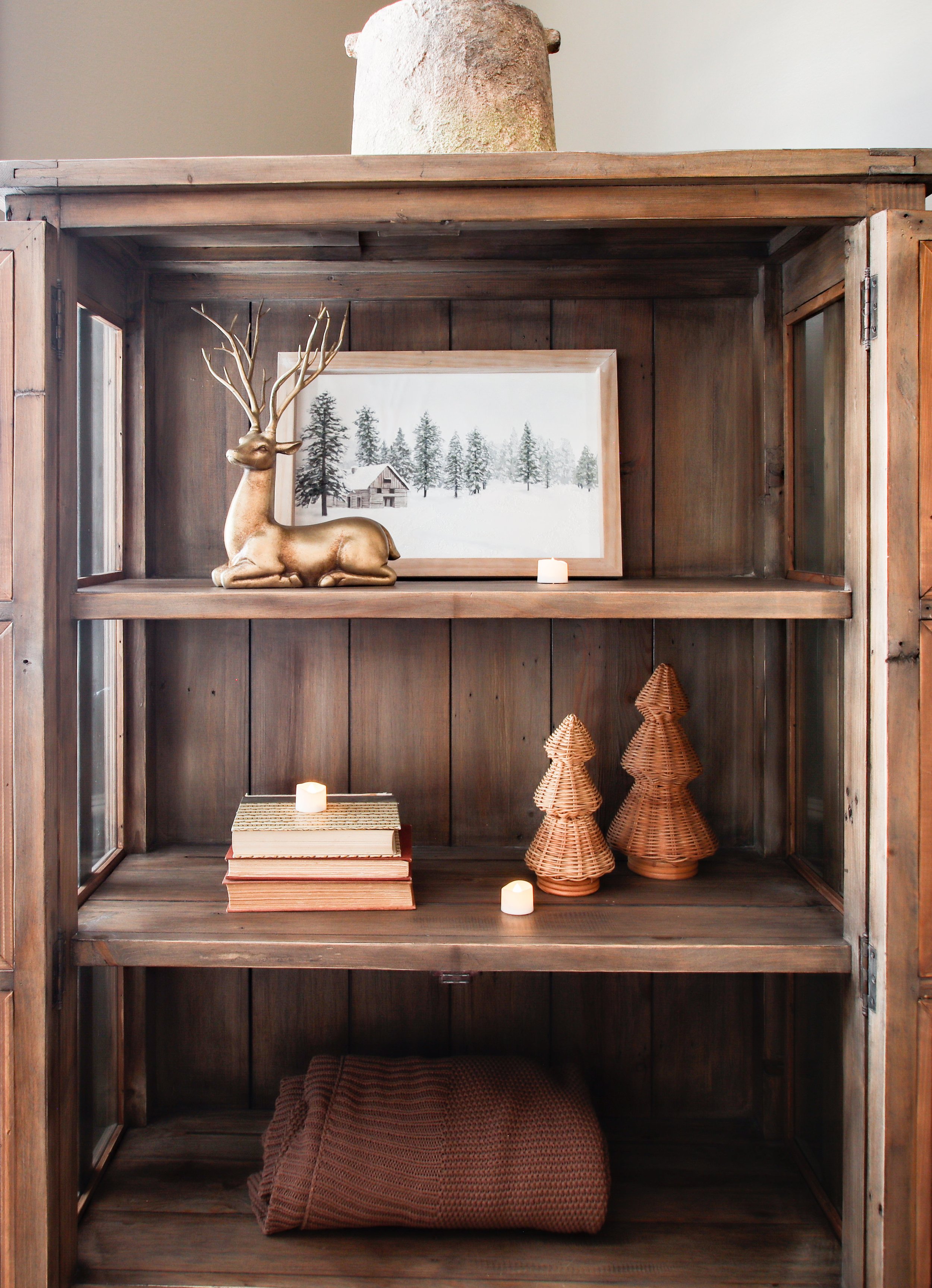 A rustic wooden shelf with decorative items including a gold-colored deer figurine, a framed winter landscape painting, small candles, woven wooden trees, and a rolled brown blanket at the bottom.