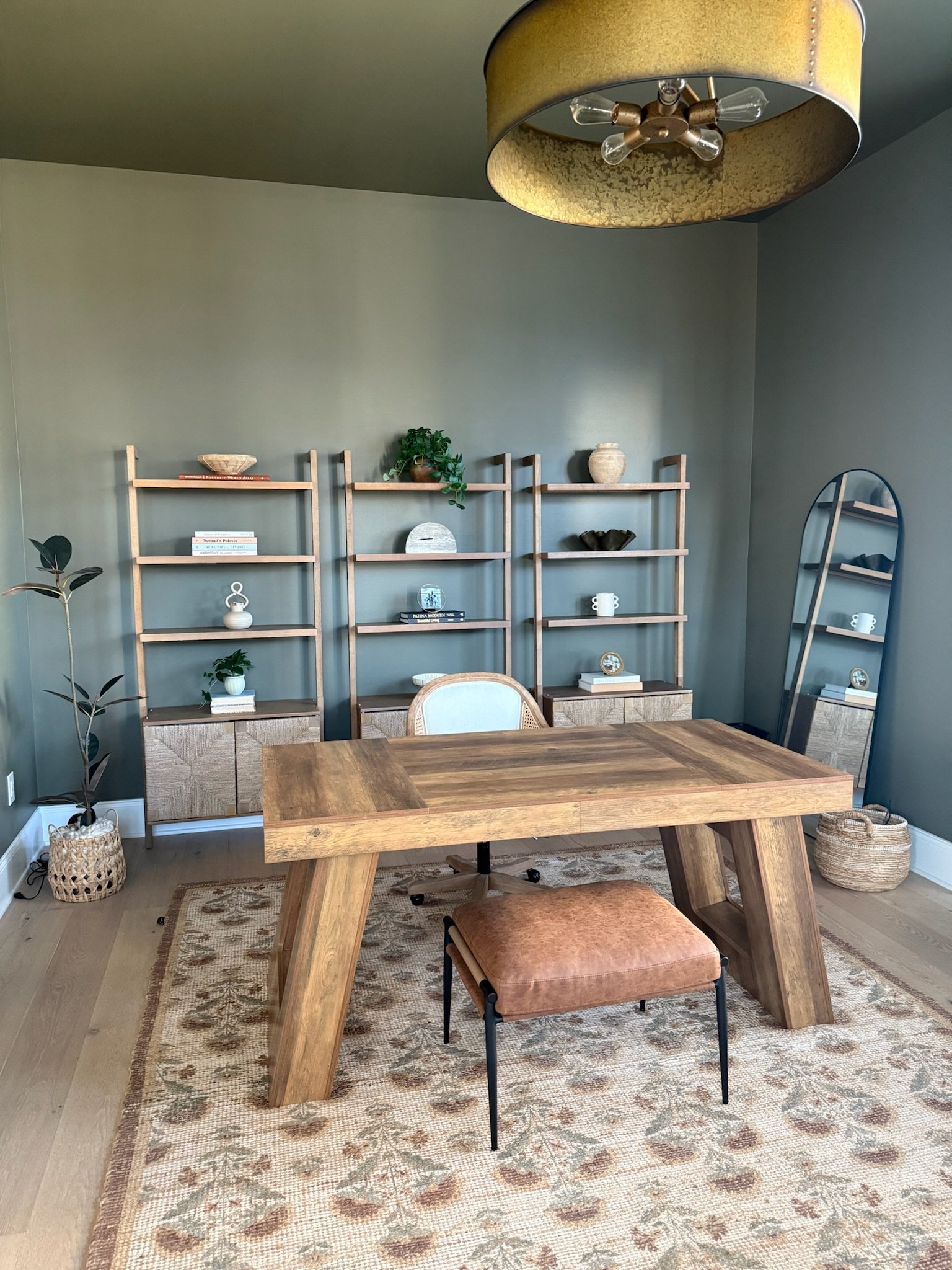 A home office with a wooden desk, beige office chair, patterned rug, a large mirror, bookshelf with decorative items, plants, and a ceiling light fixture.