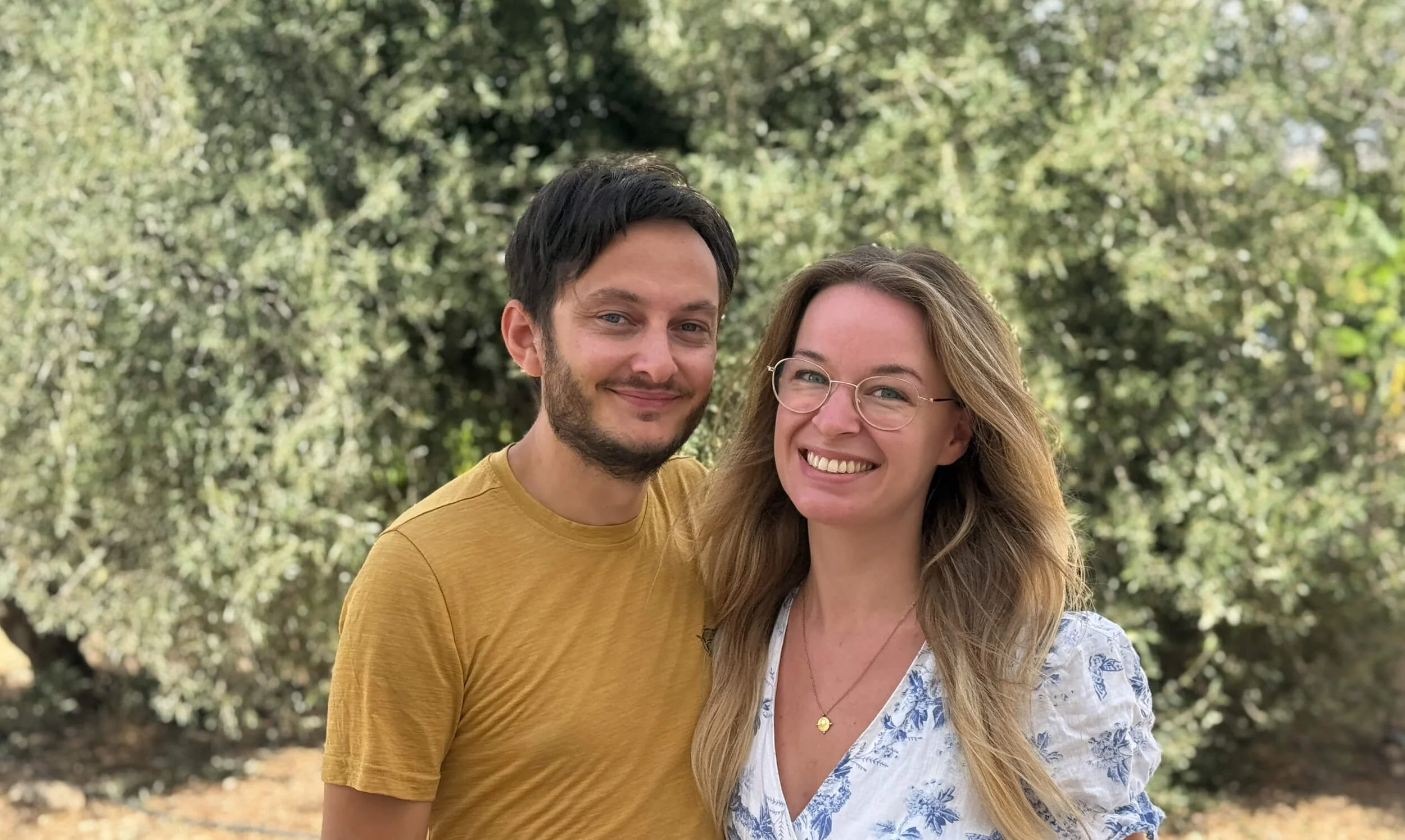 Ein lachendes Paar steht vor einem grünen, bewaldeten Hintergrund im Freien.