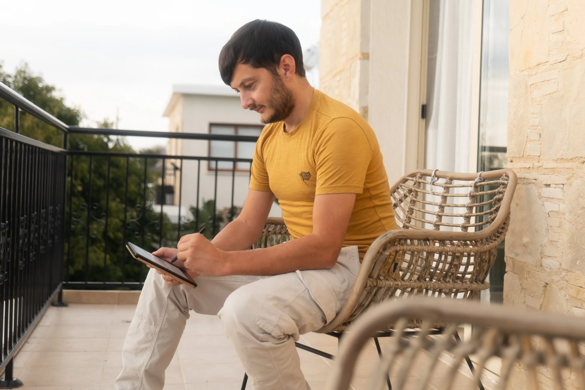 Junge Mann in gelbem T-Shirt sitzt auf einem Balkon und schreibt in ein Notizbuch.