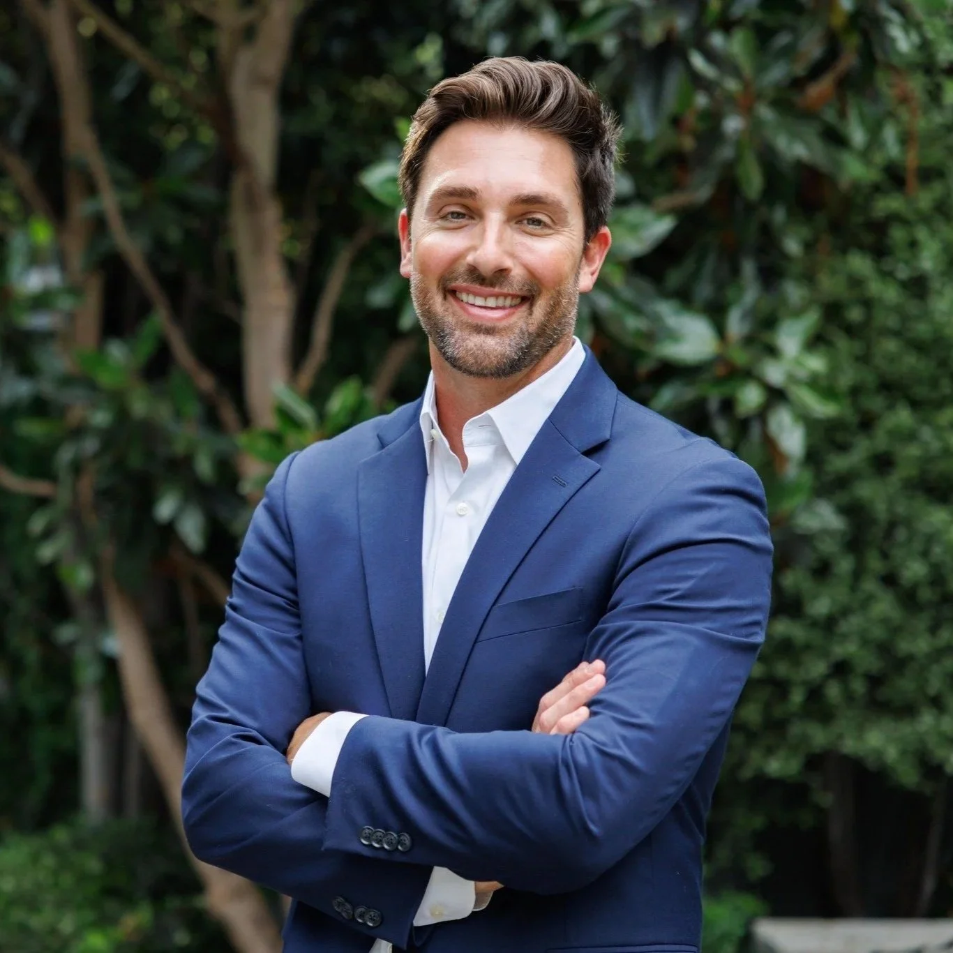 A man in a blue suit with a white shirt, smiling and standing outdoors with trees and greenery in the background.