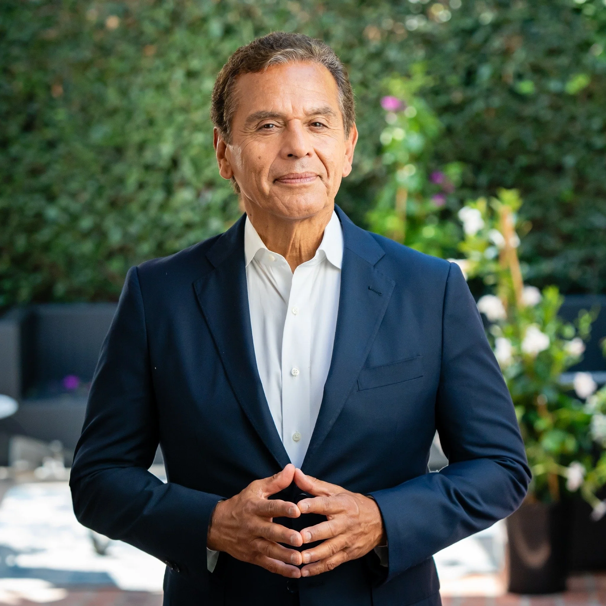 A man in a navy suit standing outdoors with a garden background.
