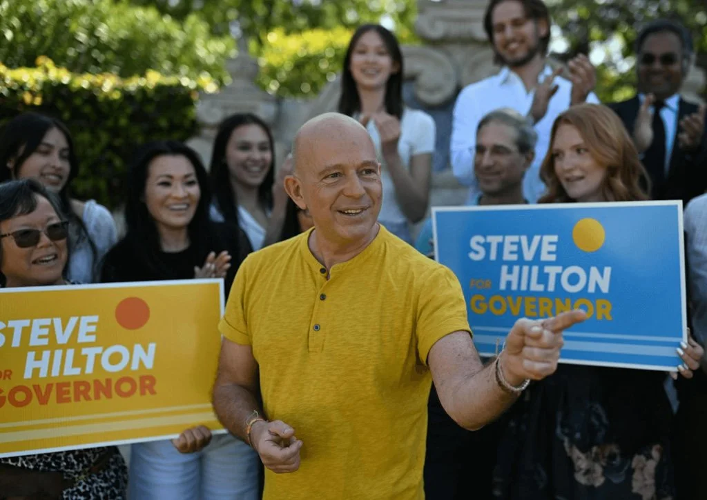 A man in a yellow shirt campaigning outdoors, holding a sign that says "Steve Hilton for Governor," surrounded by smiling supporters holding similar signs, with trees and a building in the background.