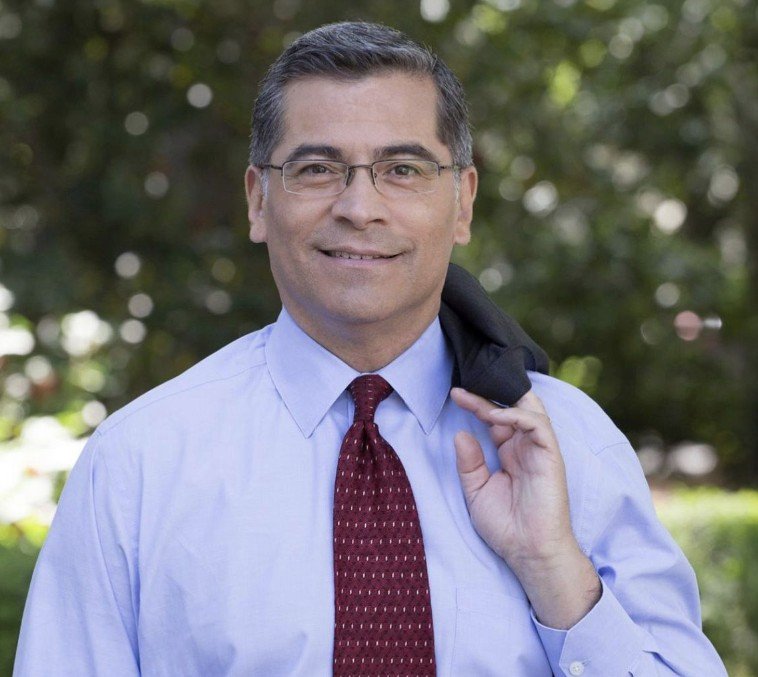 A man in glasses wearing a light blue dress shirt and a red patterned tie, holding a black jacket over his shoulder, standing outdoors with green trees in the background.