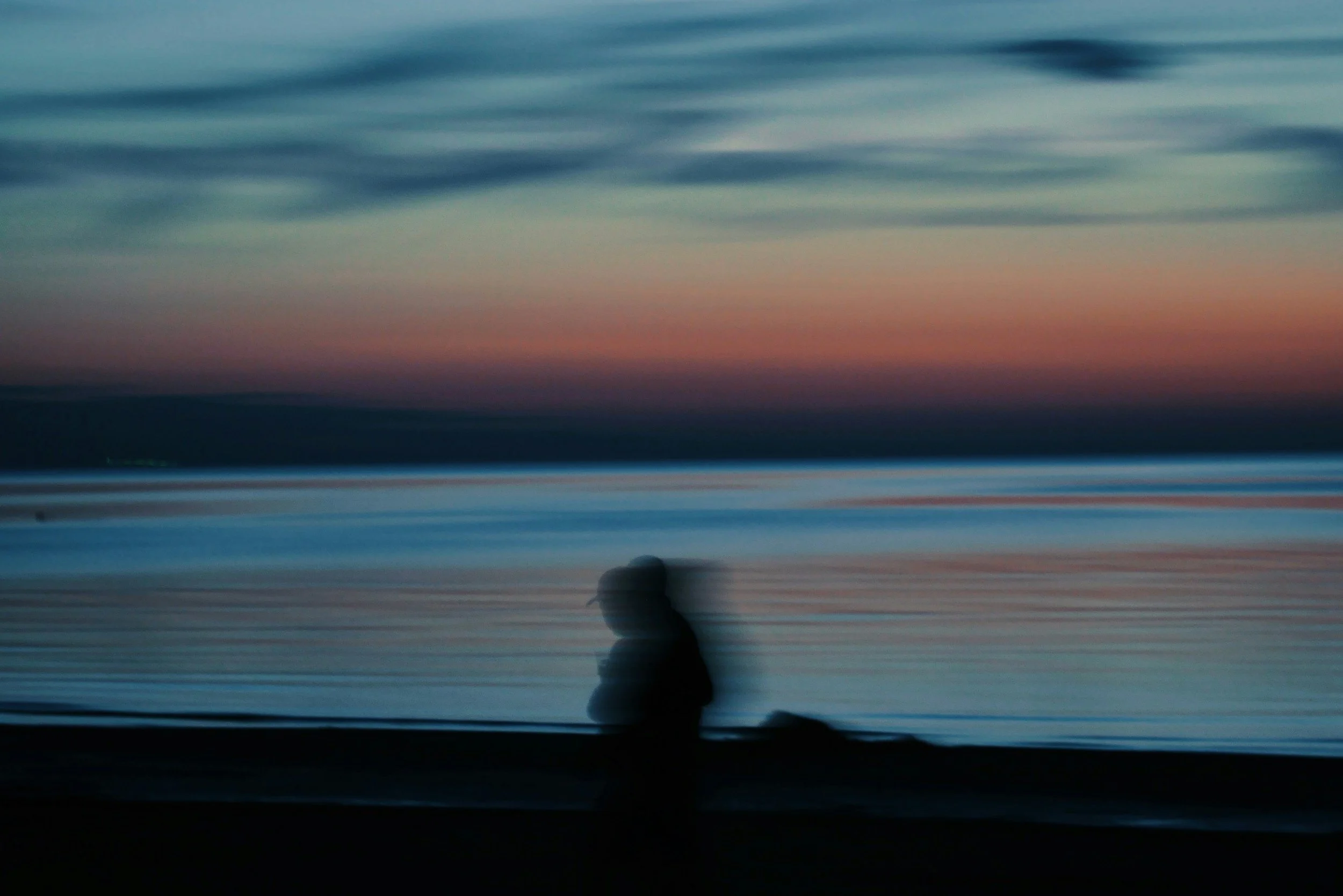 A person walking along a beach at sunset with a colorful sky and calm water.