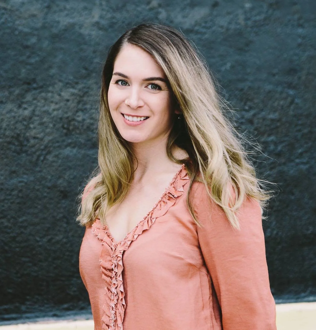 A young woman with long, wavy light brown hair and light skin, wearing a peach-colored top with ruffle details around the neckline, smiling in front of a dark, textured background.