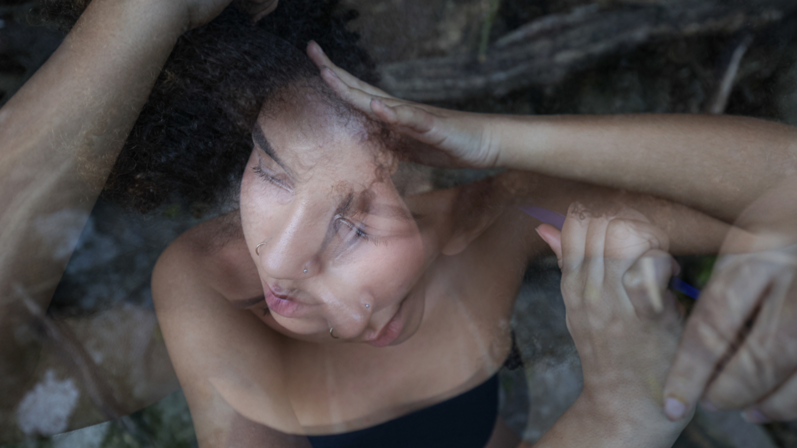 A woman with multiple facial piercings and curly hair, lying in a creek or river, with her eyes closed and hands resting on her forehead and holding something in her hand.