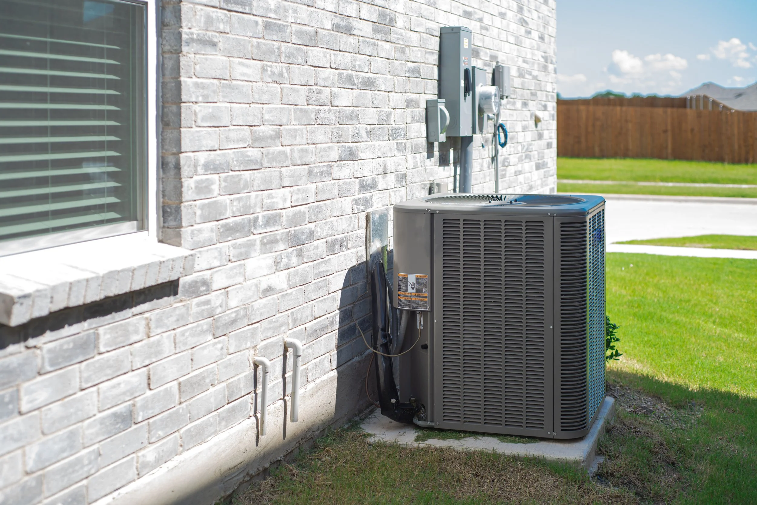 Outside view of an air conditioning unit next to a brick house wall, with electrical meters on the wall and a lawn in the background.