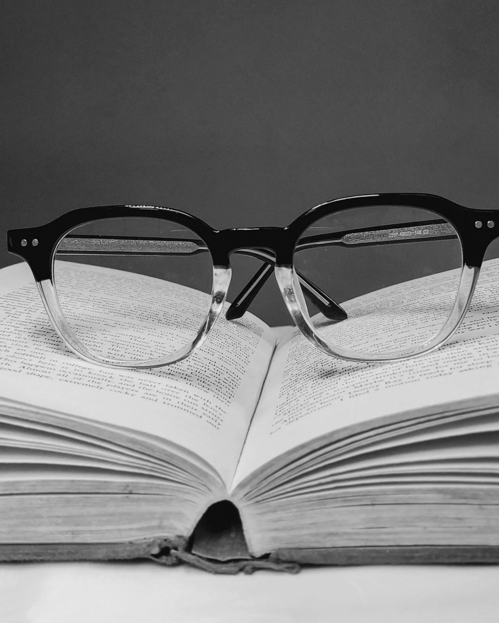 Black and clear eyeglasses resting on an open book with visible text, against a dark background.
