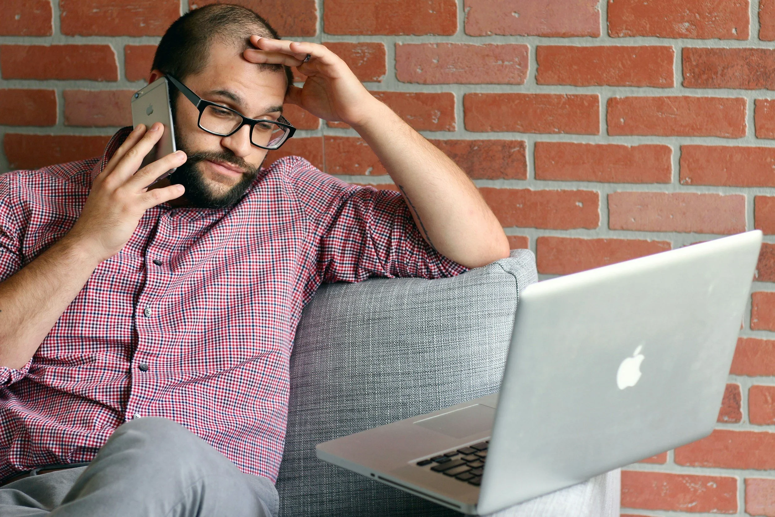 A man with glasses, a beard, and a checkered red shirt sitting on a gray sofa in front of a brick wall, holding a phone to his ear with one hand and touching his forehead with the other, with a silver MacBook on his lap.