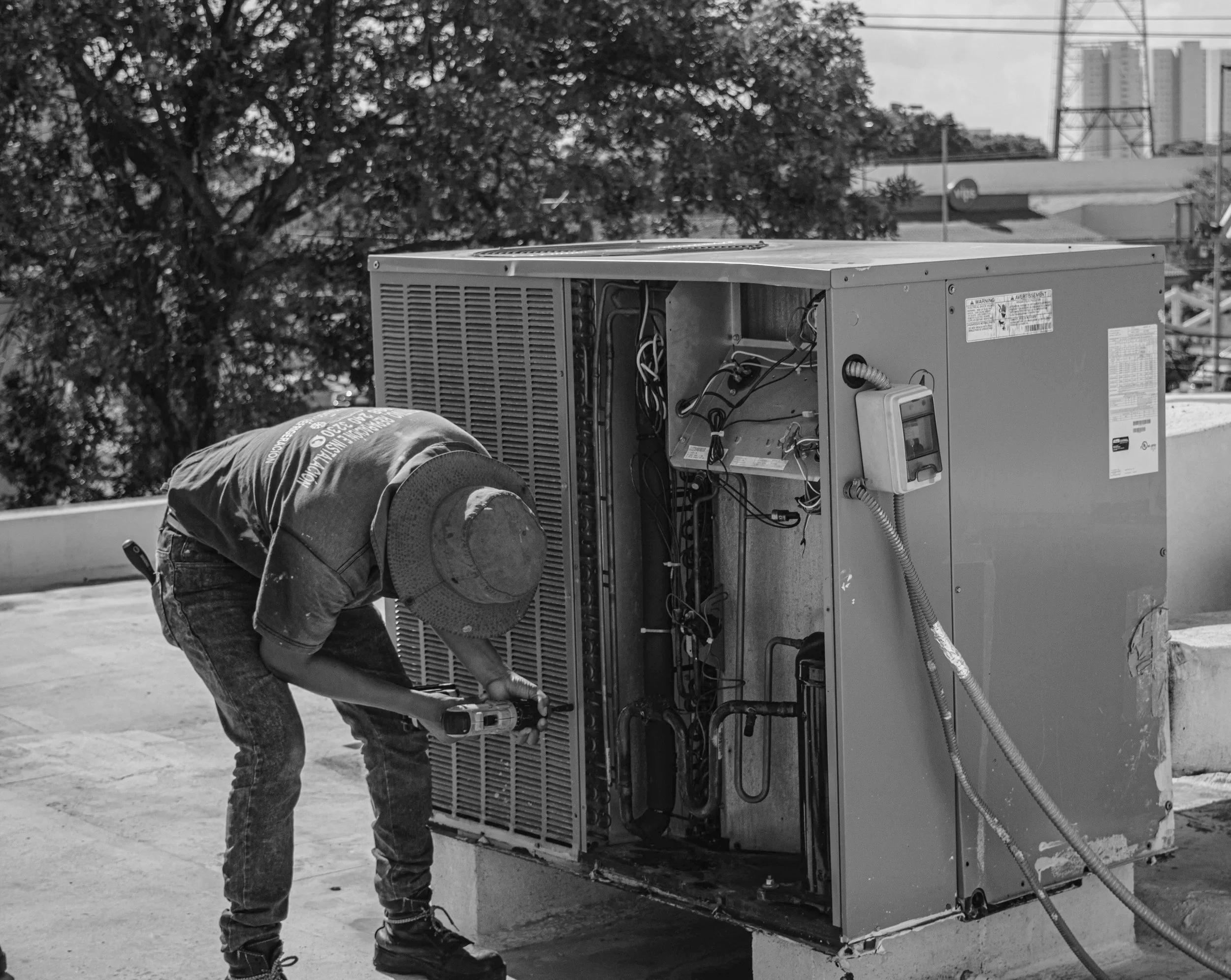 A worker wearing a hat and casual clothes is repairing or servicing an outdoor air conditioning unit on a rooftop, with the unit's cover removed to reveal internal components, during daytime.