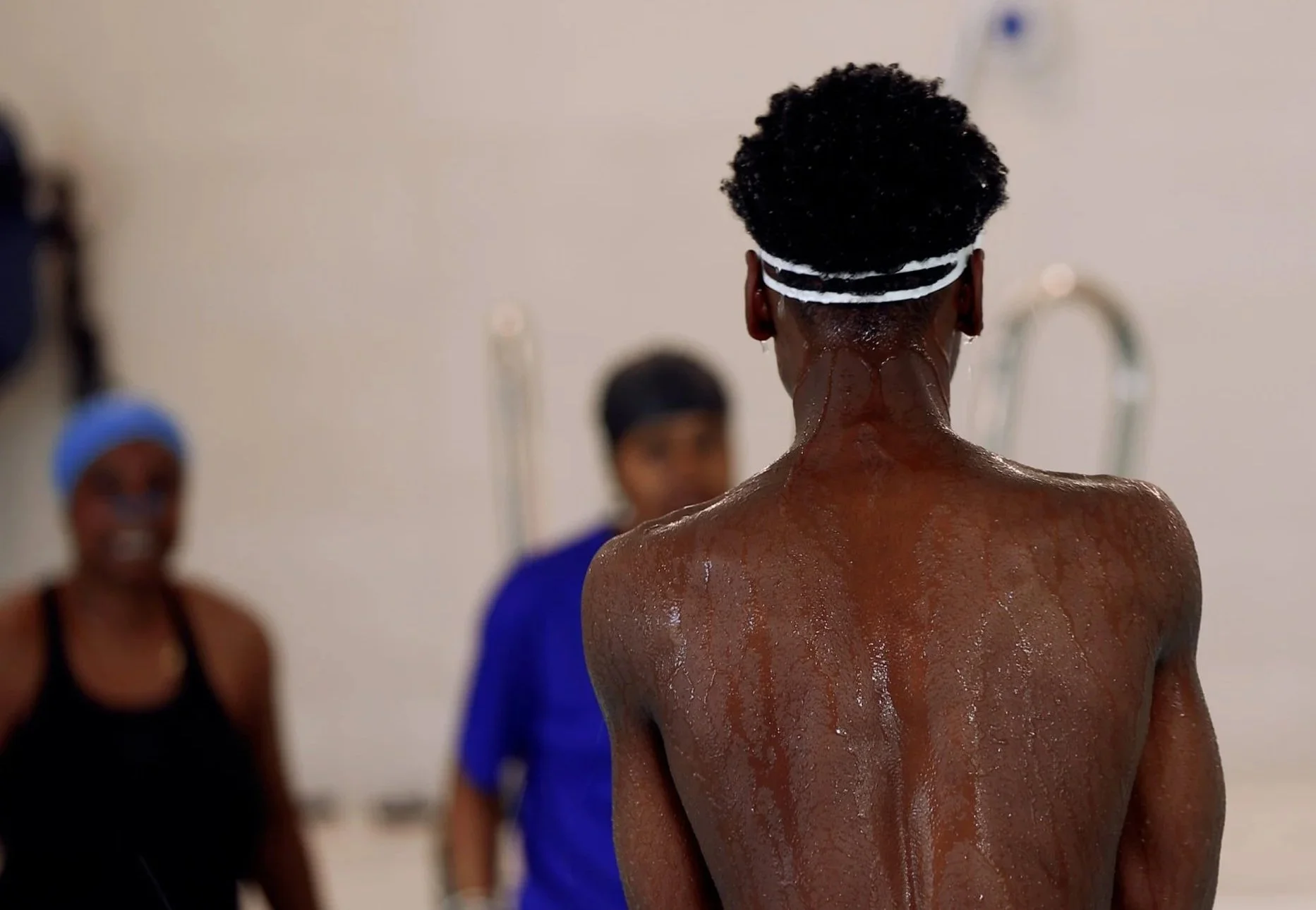 A young man with a dark Afro hairstyle wearing a white headband, shirtless, with water on his back, standing in a gym or treatment room, with two women in the background. One woman is wearing a blue cap and black tank top, and the other is dressed in blue.