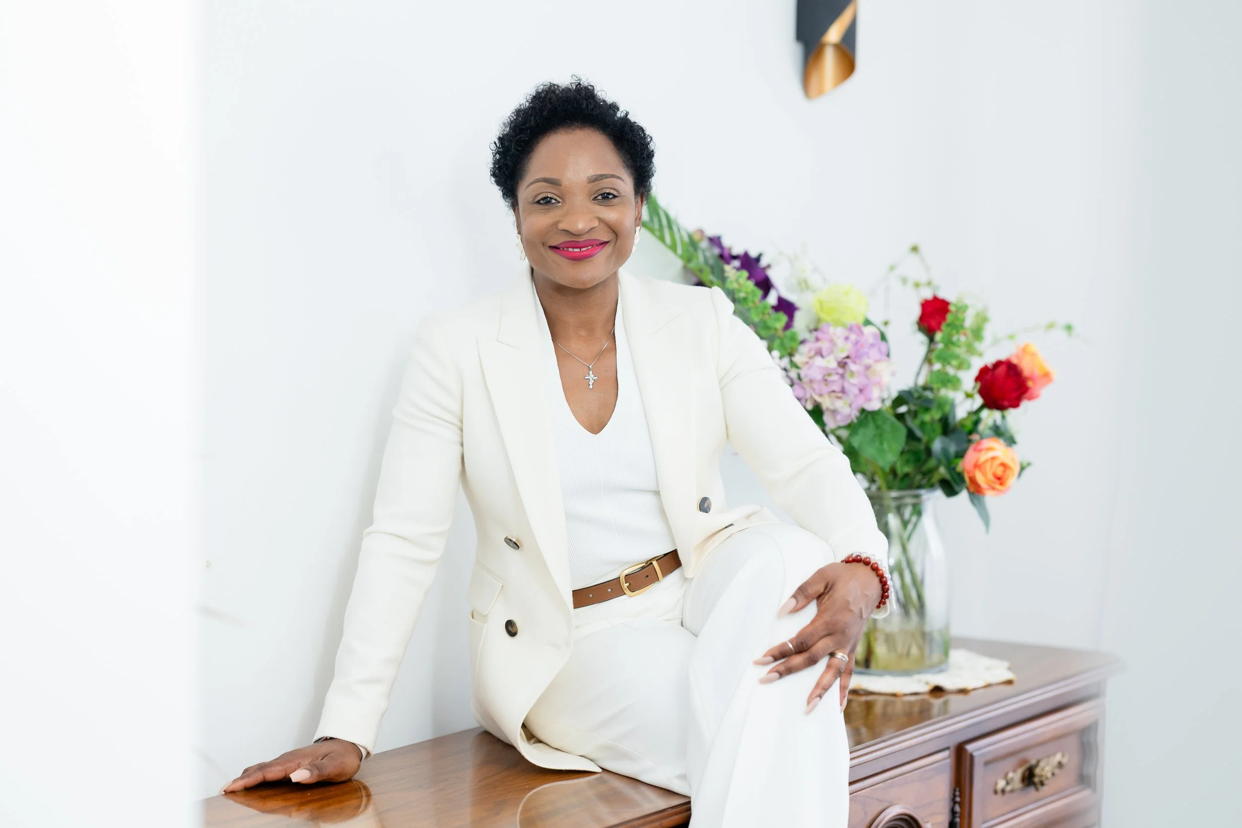 A woman in a white suit sitting on a wooden table with one leg crossed over the other, in front of a white wall with a floral arrangement in the background.