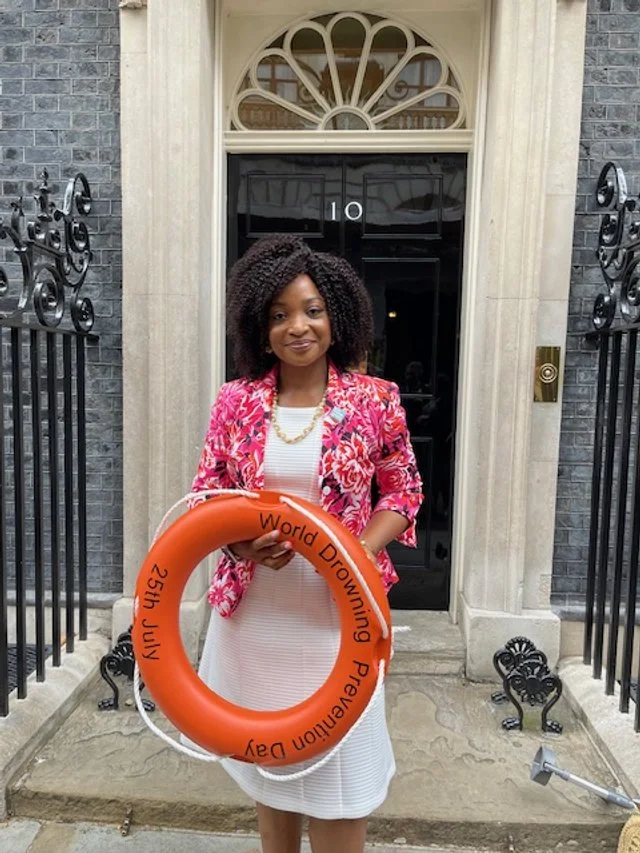 A woman standing in front of a black front door with the number 10, holding an orange life ring with the text 'World Drowning Prevention Day' and '23th July' written on it, outside a building with grey brick walls and decorative black iron railings.