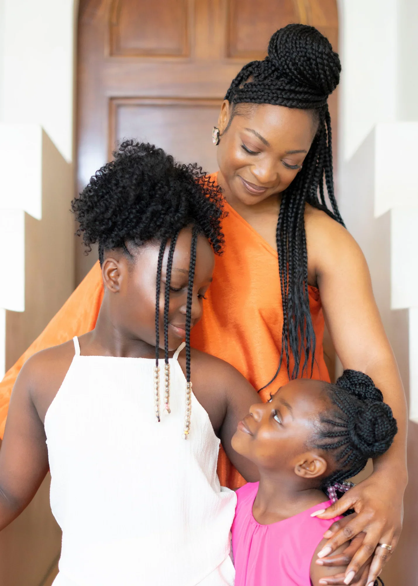 A woman with long braided hair and earrings embraces two young girls, one with curly hair and the other with braids, inside a home with a wooden door in the background.