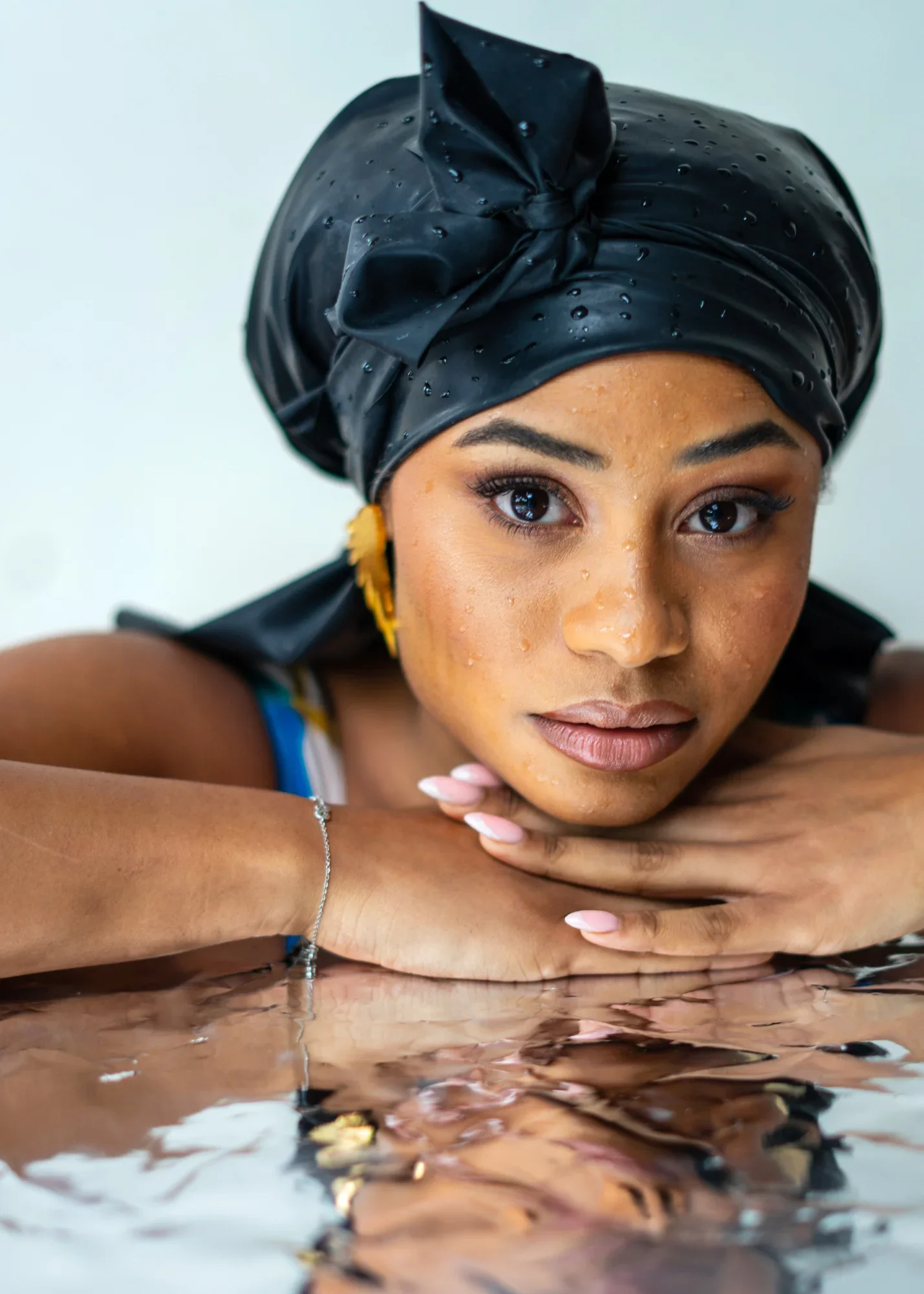A woman with water droplets on her skin, wearing a black headscarf, resting her face on her arms with reflections in water.