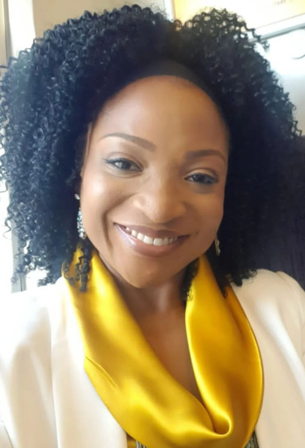 A woman smiling with curly black hair, wearing earrings, a white blazer, and a yellow satin scarf, indoors with a blurred background.