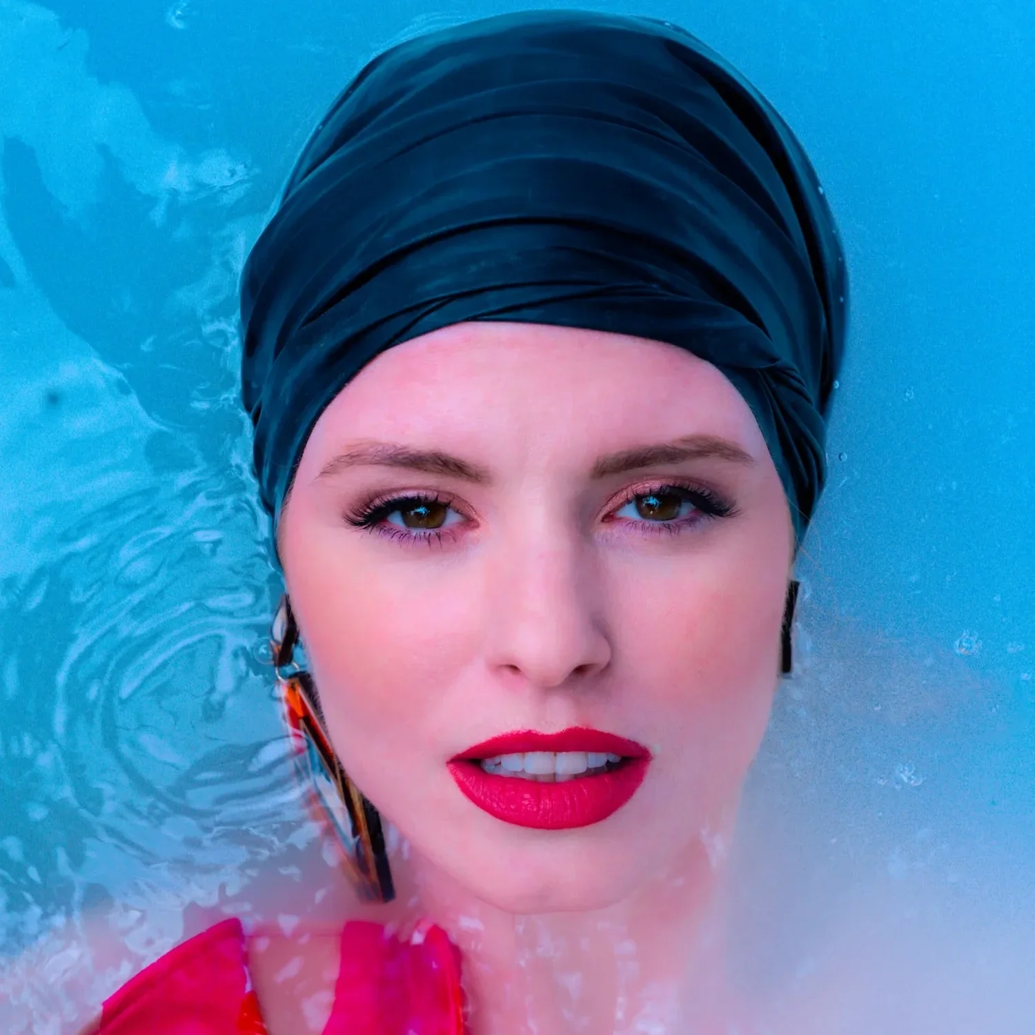 A woman with makeup and red lipstick wearing a black swim cap, partially submerged in water, looking at the camera.