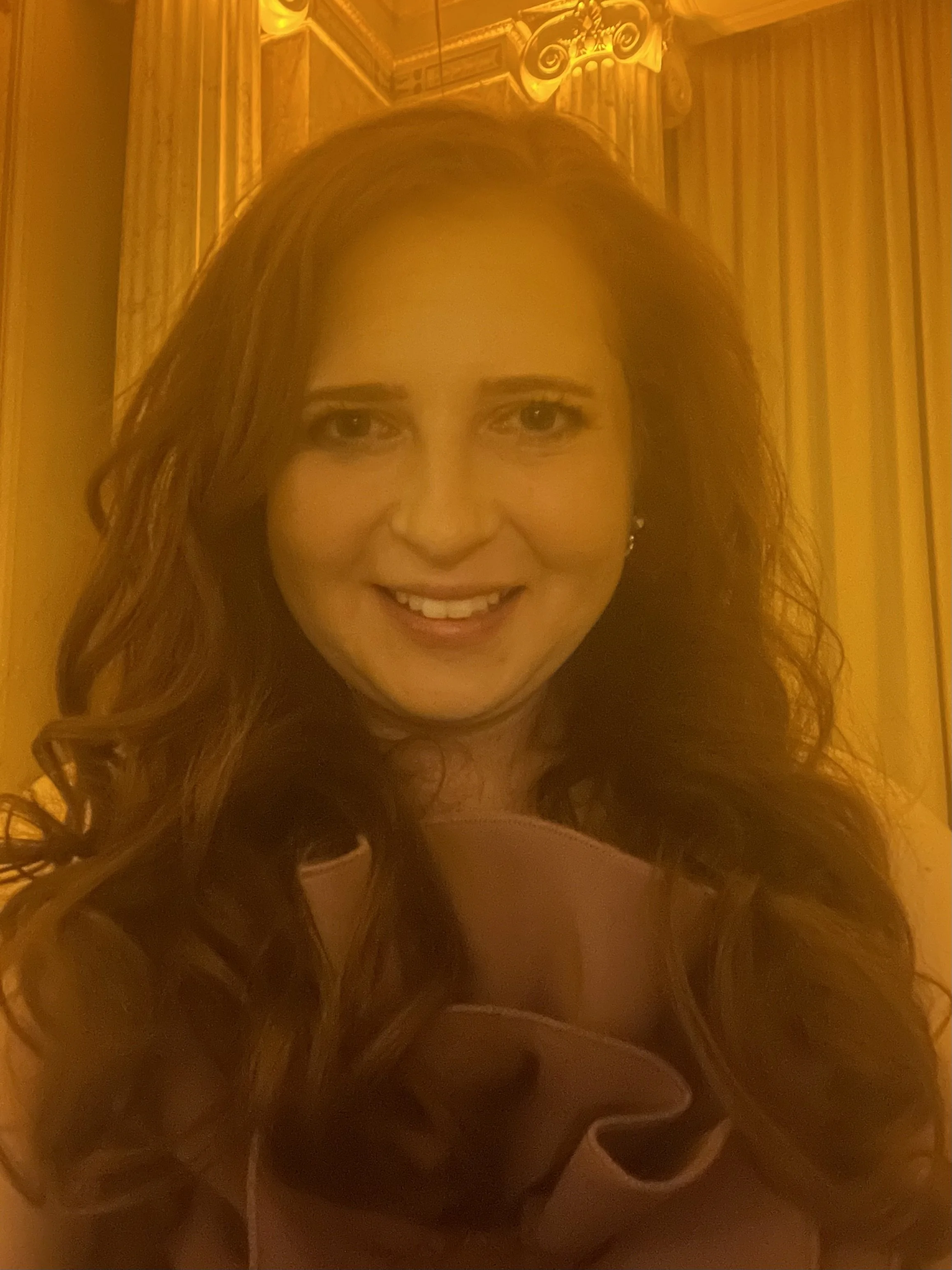 A woman with long, wavy brown hair smiling at the camera in an elegant room with gold accents and curtains.